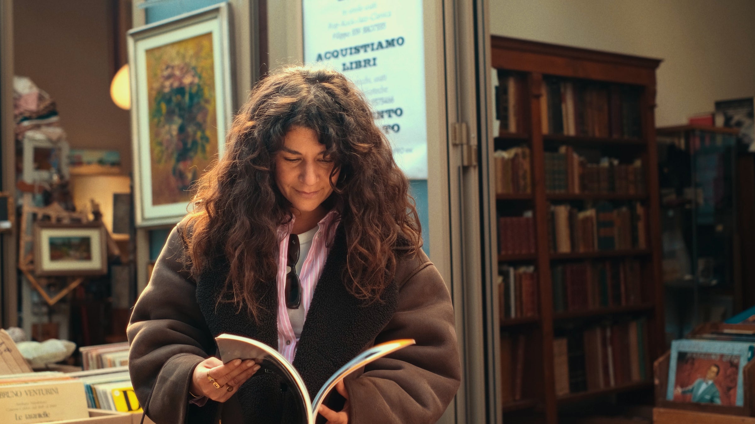 A woman with long curly hair and a brown coat stands in a cozy bookstore, smiling as she reads an open book. Bookshelves and framed art are visible in the warmly lit background.
