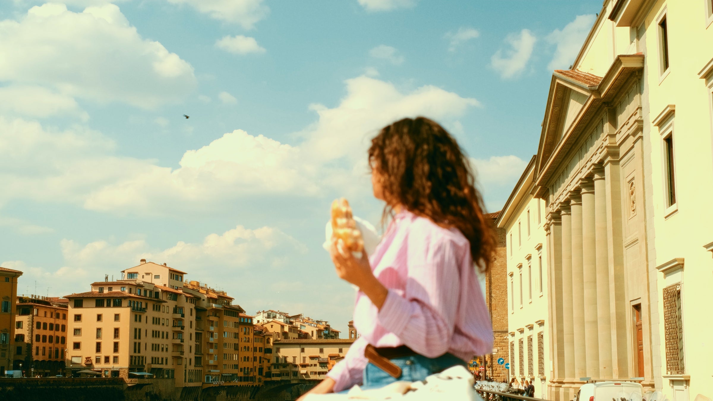 A person with curly hair, wearing a pink shirt, holds food while sitting outdoors. Historic buildings and a blue sky with clouds are in the background. The focus is on the scenery, with the person slightly blurred.