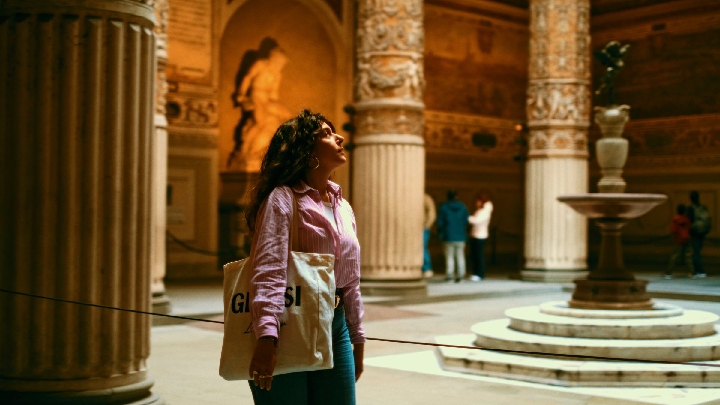 A woman with curly hair, wearing a pink shirt and carrying a tote bag, stands in a historic, ornately decorated hall with columns and statues, looking up at the artwork on the walls.