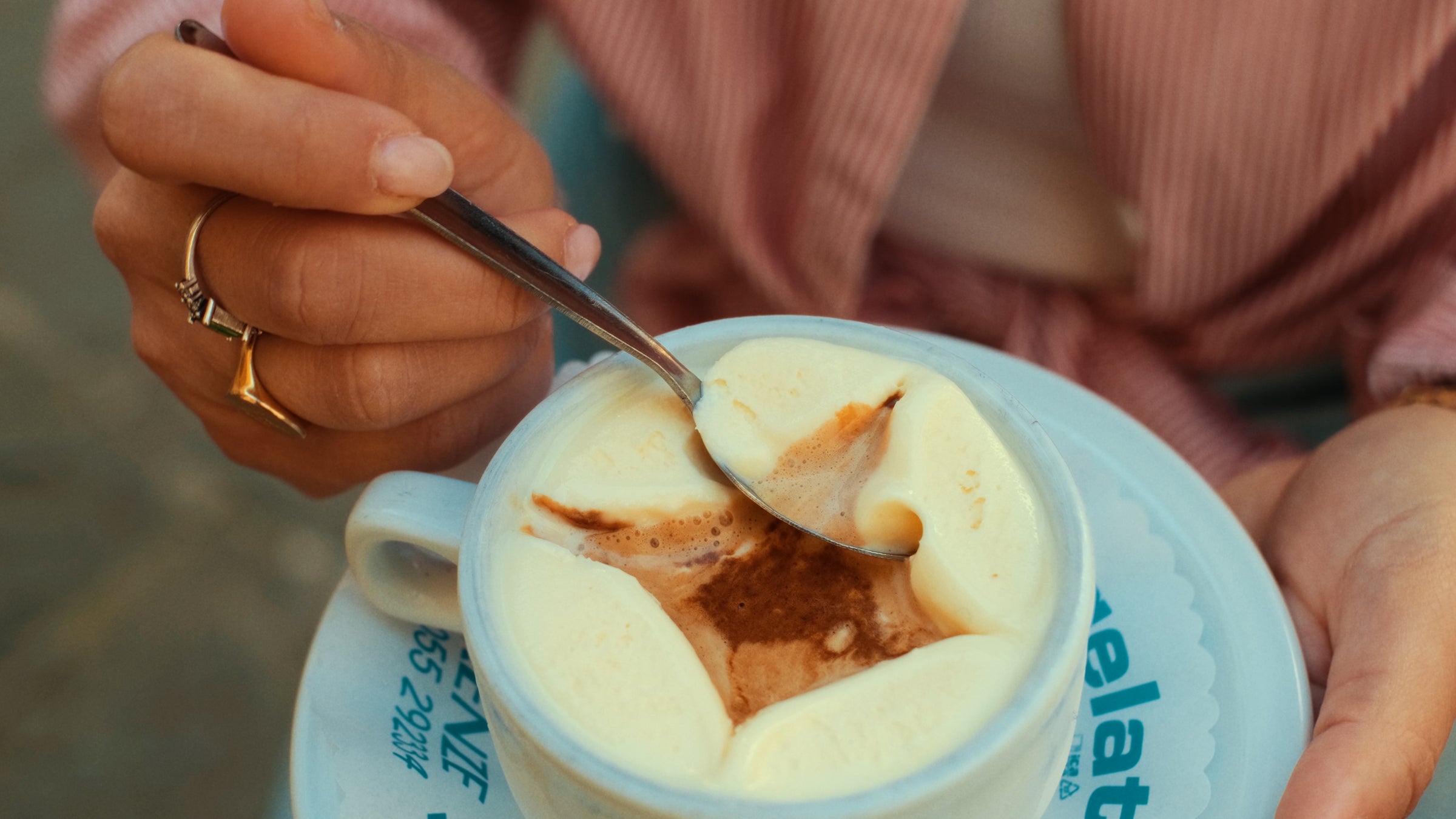A person holding a spoon stirs a cup of coffee topped with creamy foam, sitting on a white saucer. The person is wearing rings and a pink striped shirt.