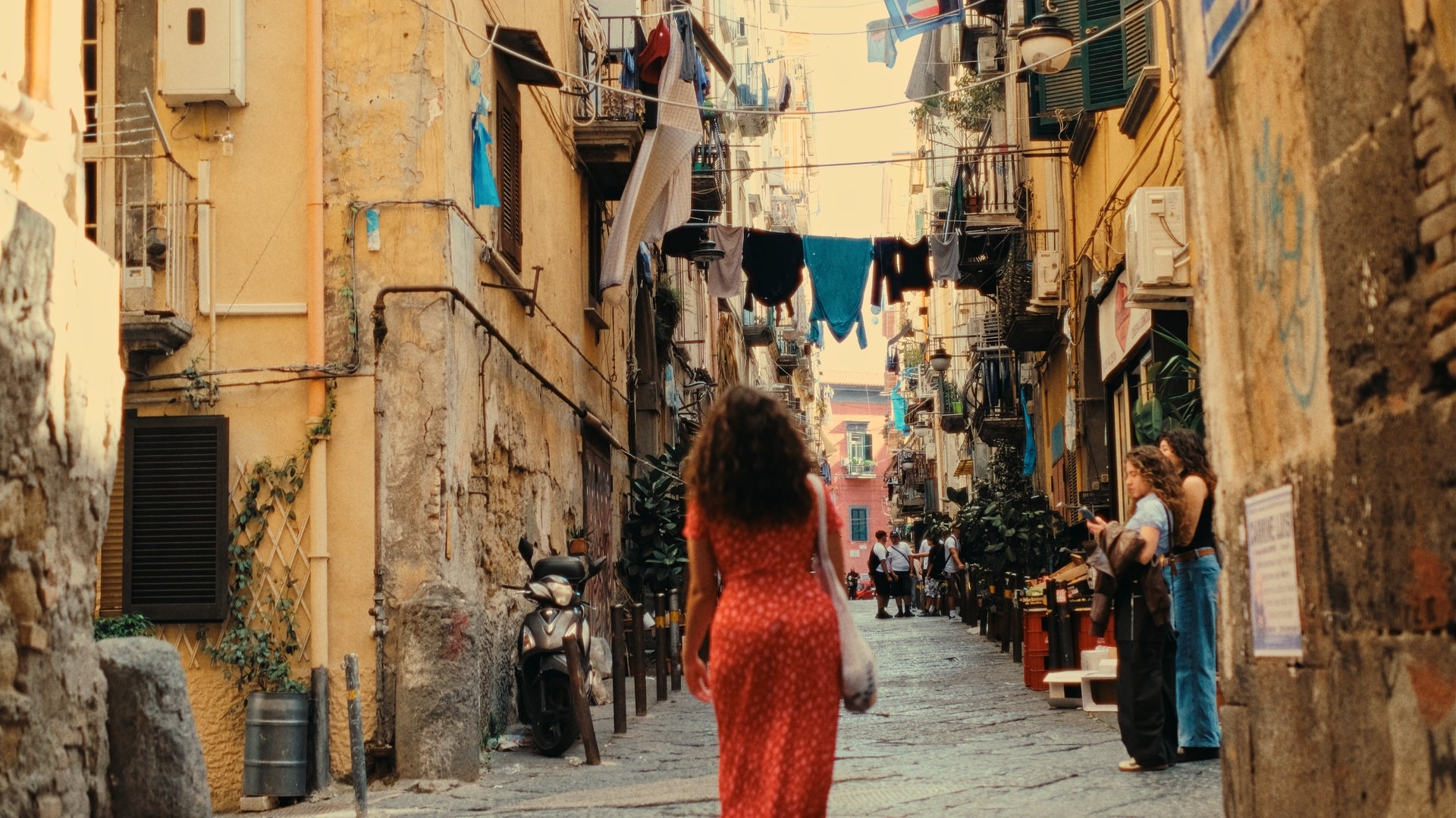 A woman in a red dress walks down a narrow, sunlit street lined with old buildings, laundry hanging on clotheslines above, and people chatting along the sides.