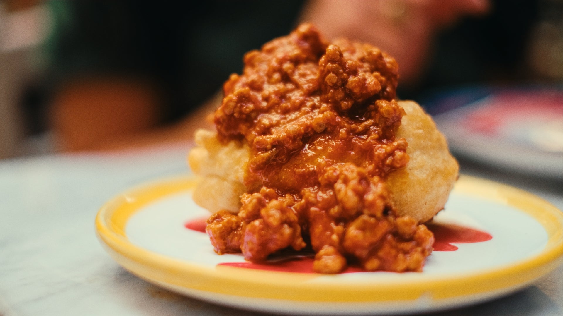 A close-up of a pastry topped with a generous serving of saucy ground meat, served on a white plate with a yellow rim, on a table with a blurred background.