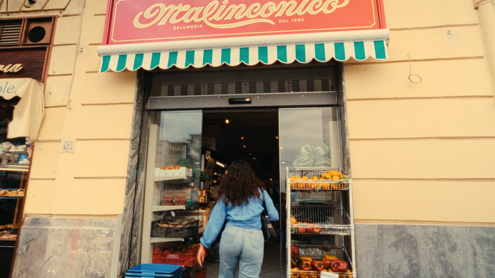 A woman with long, curly hair and wearing light blue clothing enters a small grocery store with striped green and white awning and red sign above the entrance. Fresh produce is displayed in bins outside the door.
