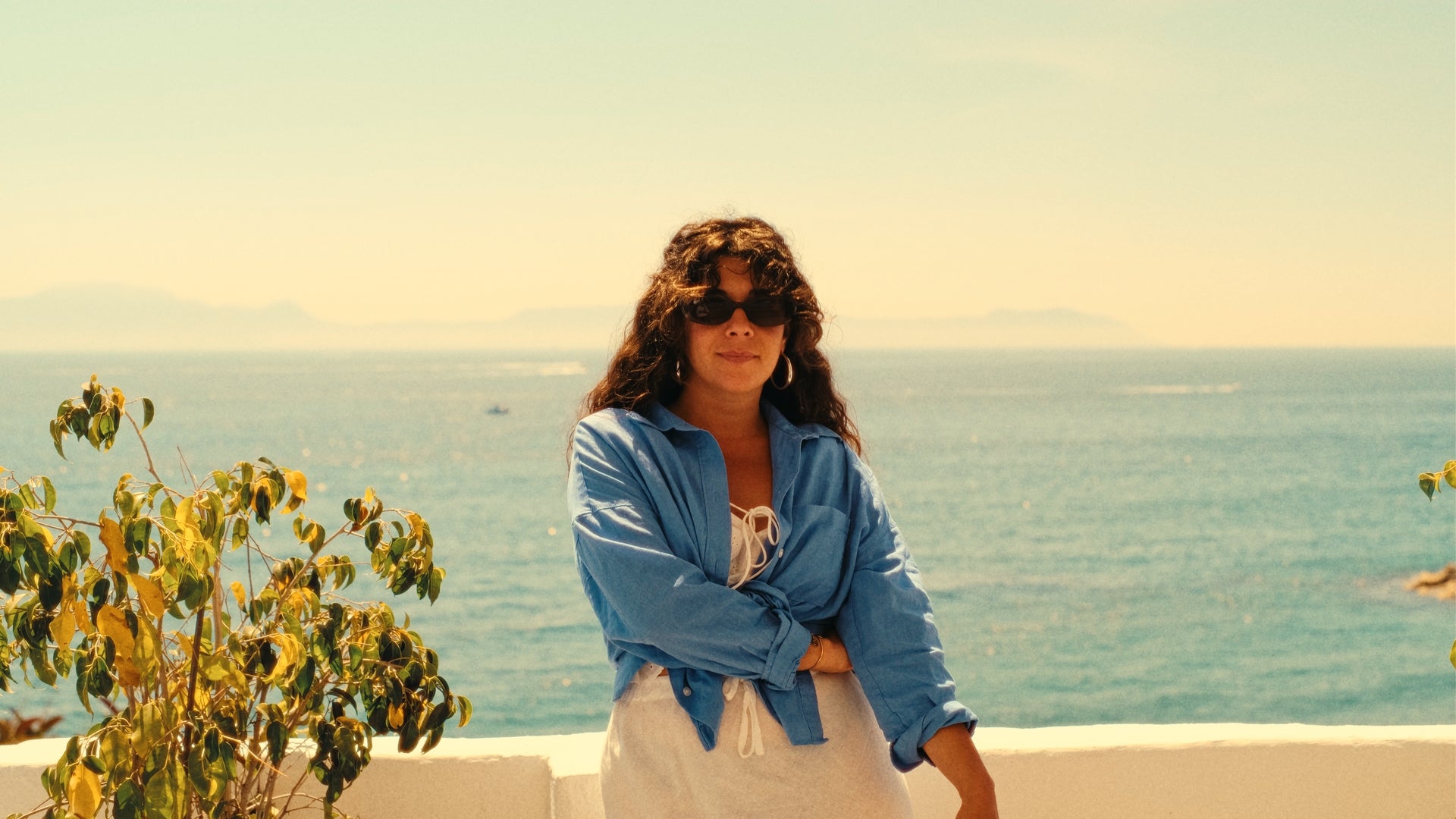 A woman with curly hair and sunglasses stands by a white wall, wearing a blue shirt over a white outfit. The background shows the ocean, a clear sky, and distant mountains on a sunny day.