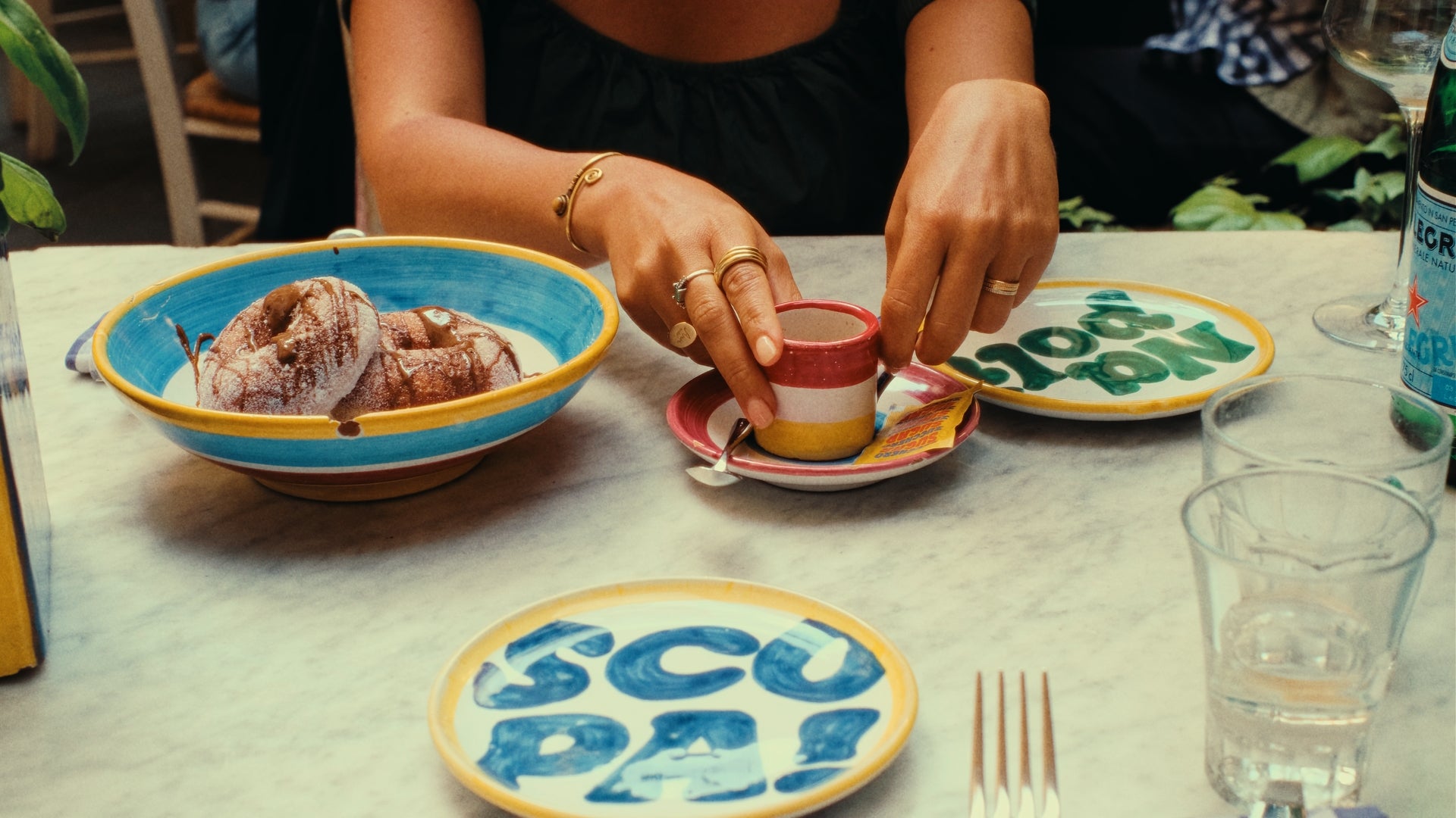 A person with rings on their fingers prepares espresso at a table set with colorful plates, a bowl of pastries, a water bottle, and drinking glasses on a marble surface.