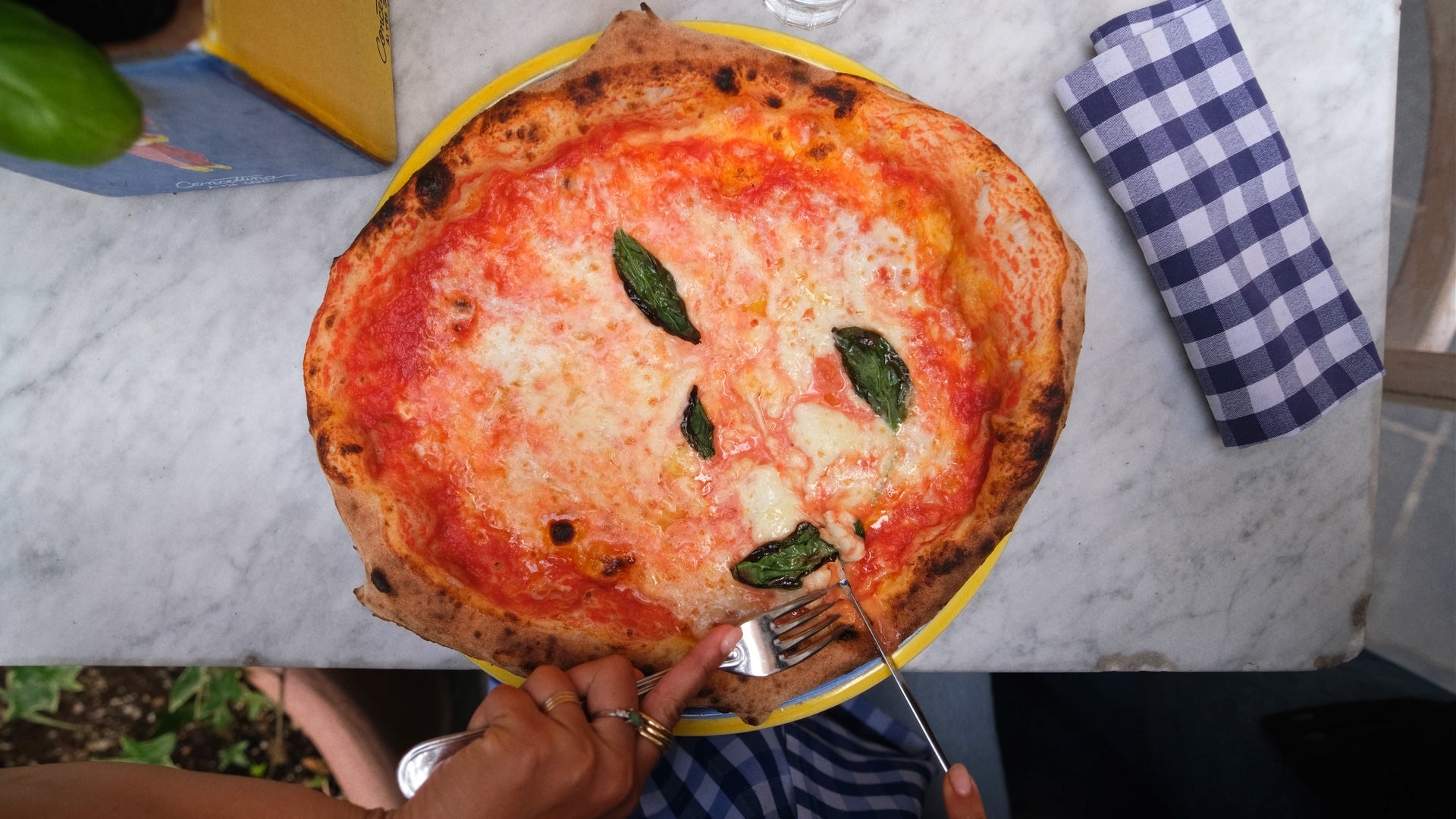 A hand holding a fork and knife cuts into a Margherita pizza topped with mozzarella and basil leaves on a yellow plate, placed on a white marble table beside a blue checkered napkin.
