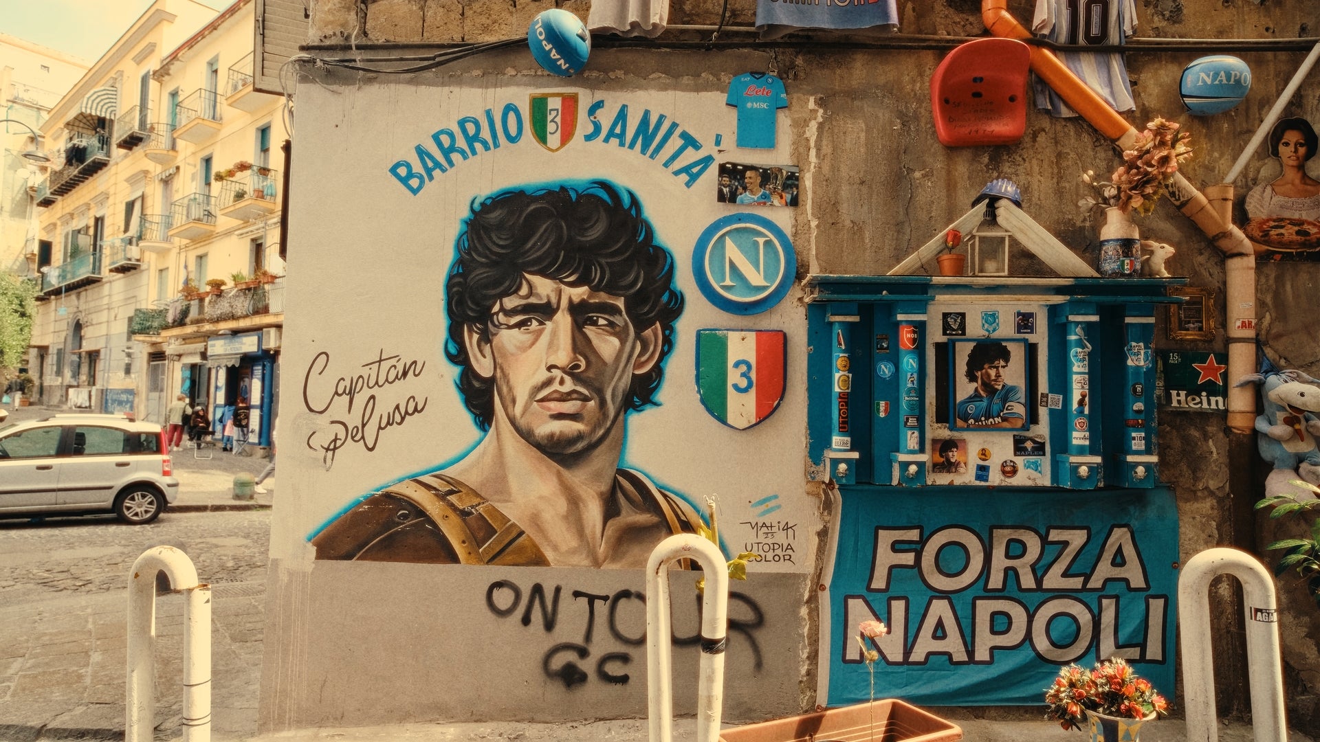 A colorful street mural in Naples features a portrait of Diego Maradona, surrounded by Napoli football memorabilia, graffiti, and the phrases Barrio Sanità and Forza Napoli. The scene is vibrant and full of local character.