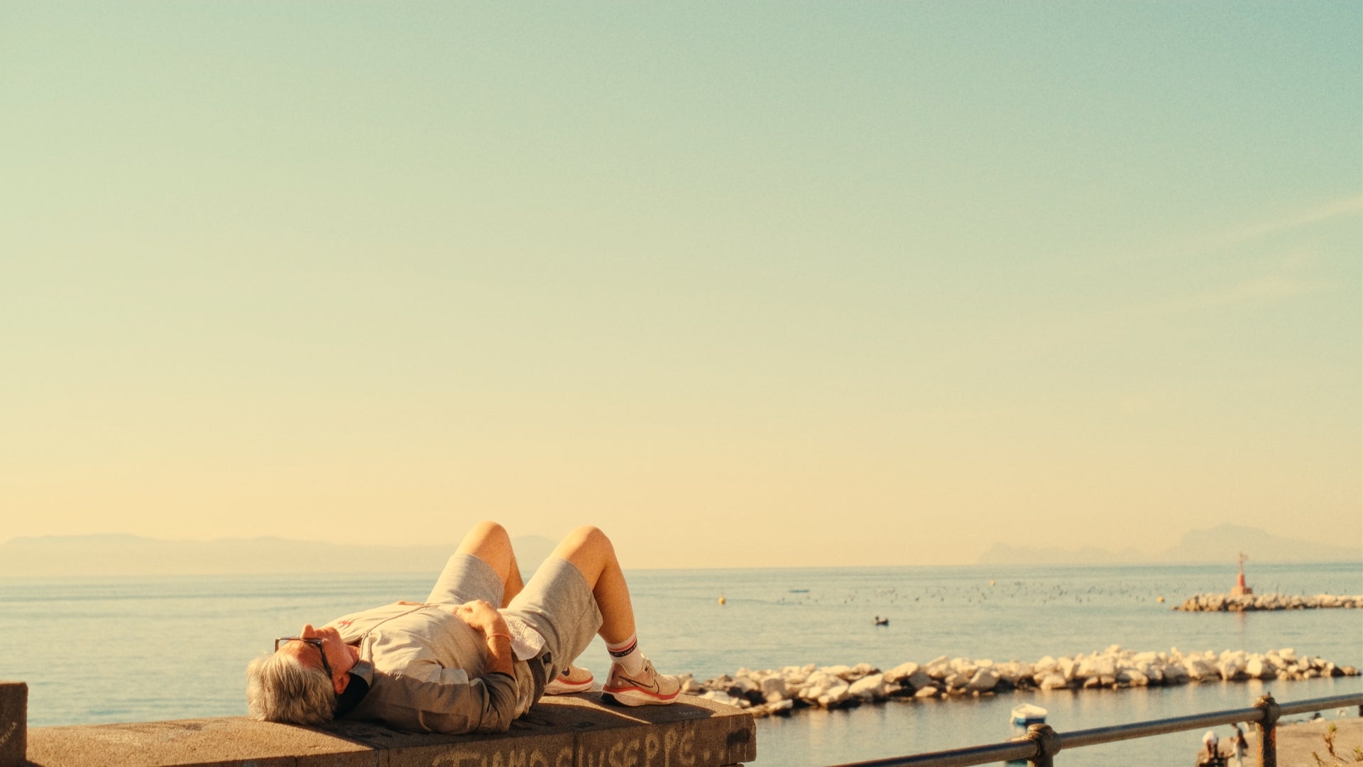 An older man in shorts and a t-shirt lies on his back on a concrete ledge by the sea, basking in the sunlight. The sky is clear and the water is calm, with distant mountains visible on the horizon.