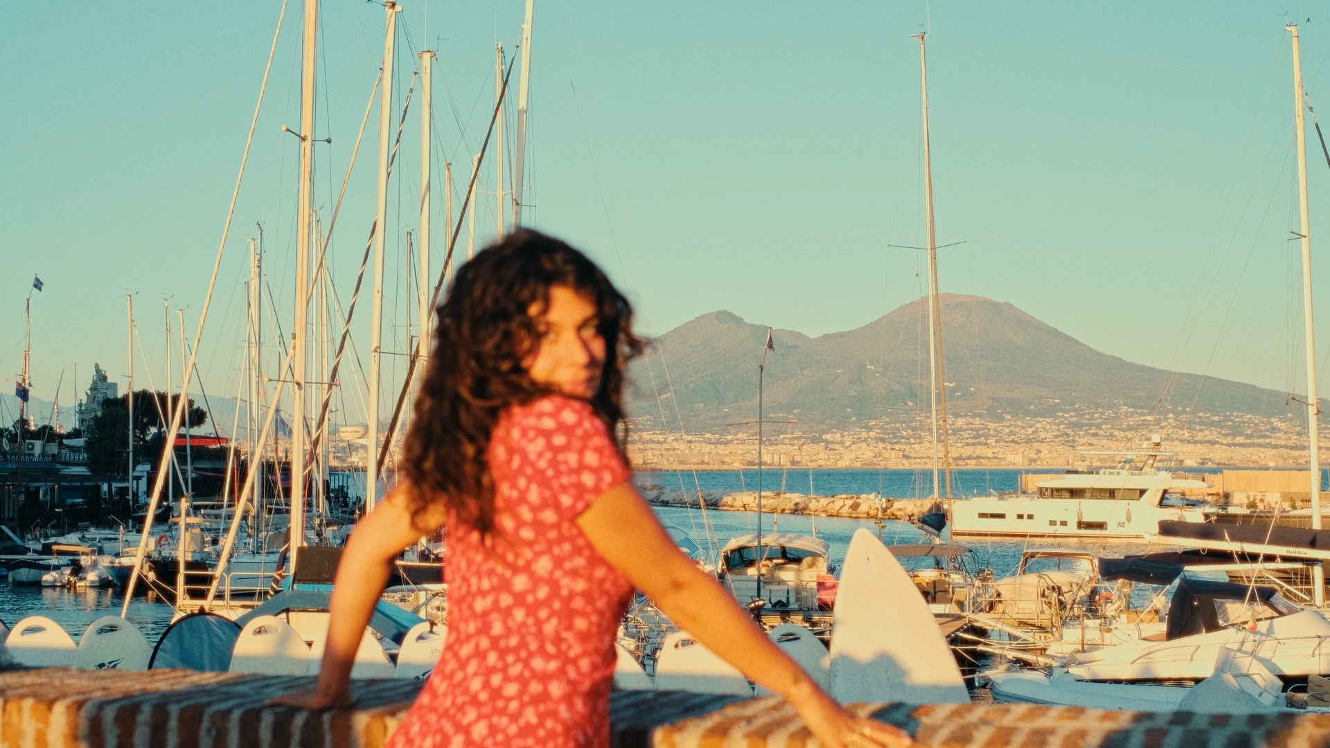 A woman in a red dress with curly hair leans on a stone railing, with a marina full of boats and sailboats behind her. Distant mountains and a clear blue sky complete the scenic background.