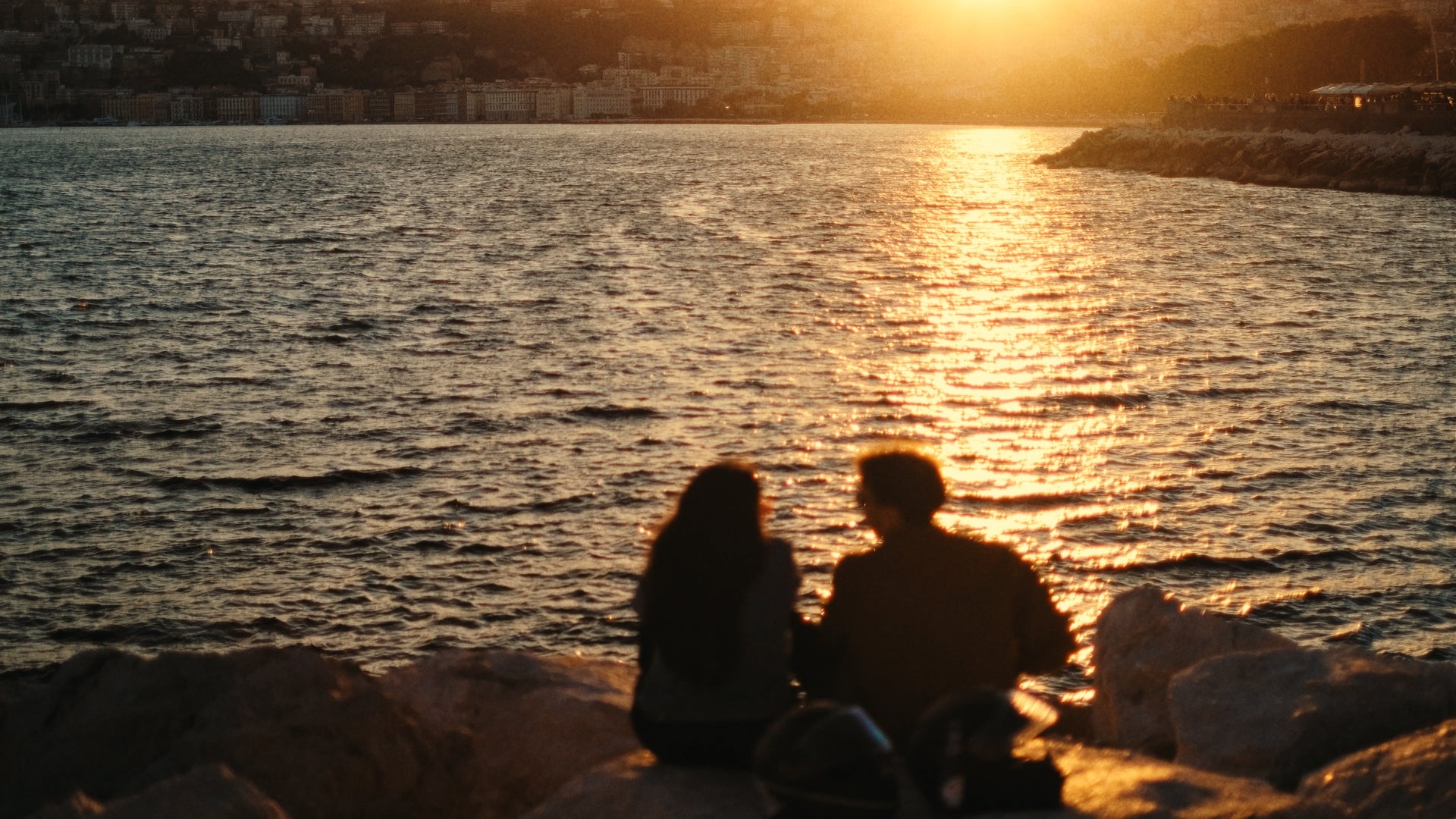 Two people sit on rocks by the water, silhouetted against a golden sunset. The sunlight reflects off the waves, with a cityscape and hills visible in the background.