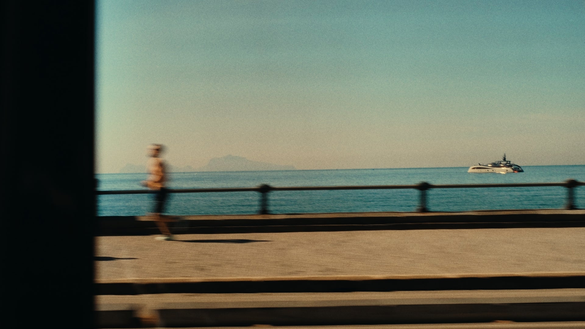 A blurry person jogs along a seaside promenade under a clear sky, with a large yacht visible on the calm blue water in the background. A faint outline of distant mountains is seen on the horizon.