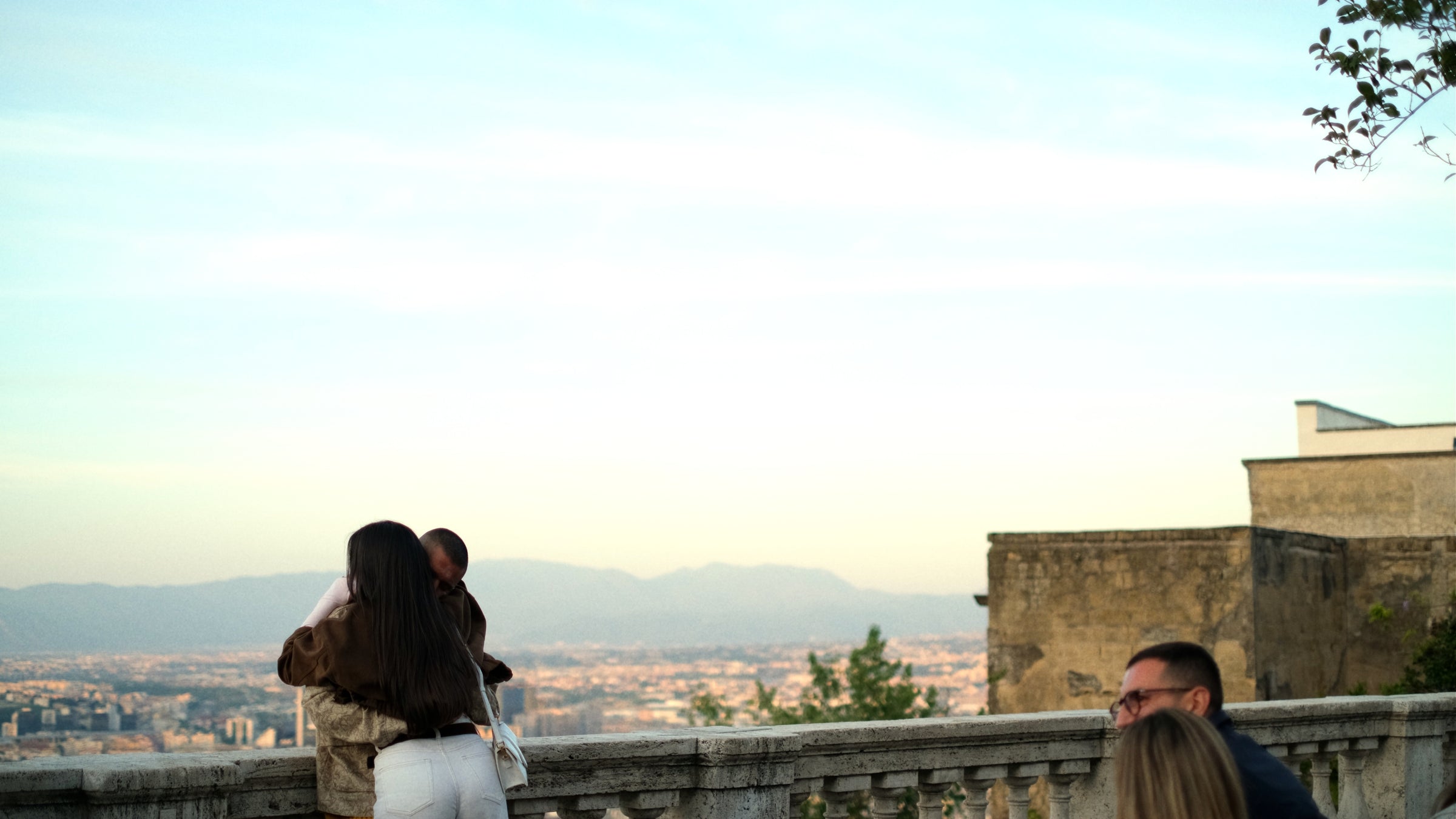 A woman holding a baby stands by a stone railing, overlooking a cityscape with mountains in the distance. Two other people are seated nearby, partially visible in the foreground. The sky is clear and pale blue.