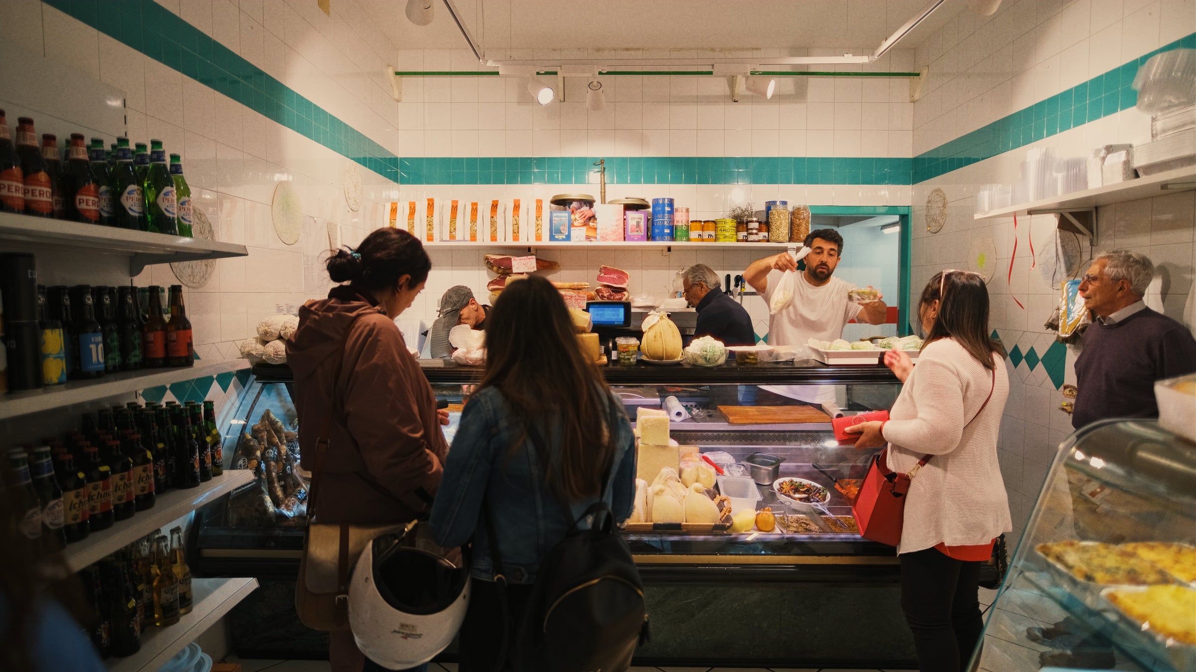 Several people stand inside a small deli with tiled walls, browsing and ordering food from a display counter filled with cheeses and deli products. A man behind the counter serves customers. Shelves hold drinks and groceries.