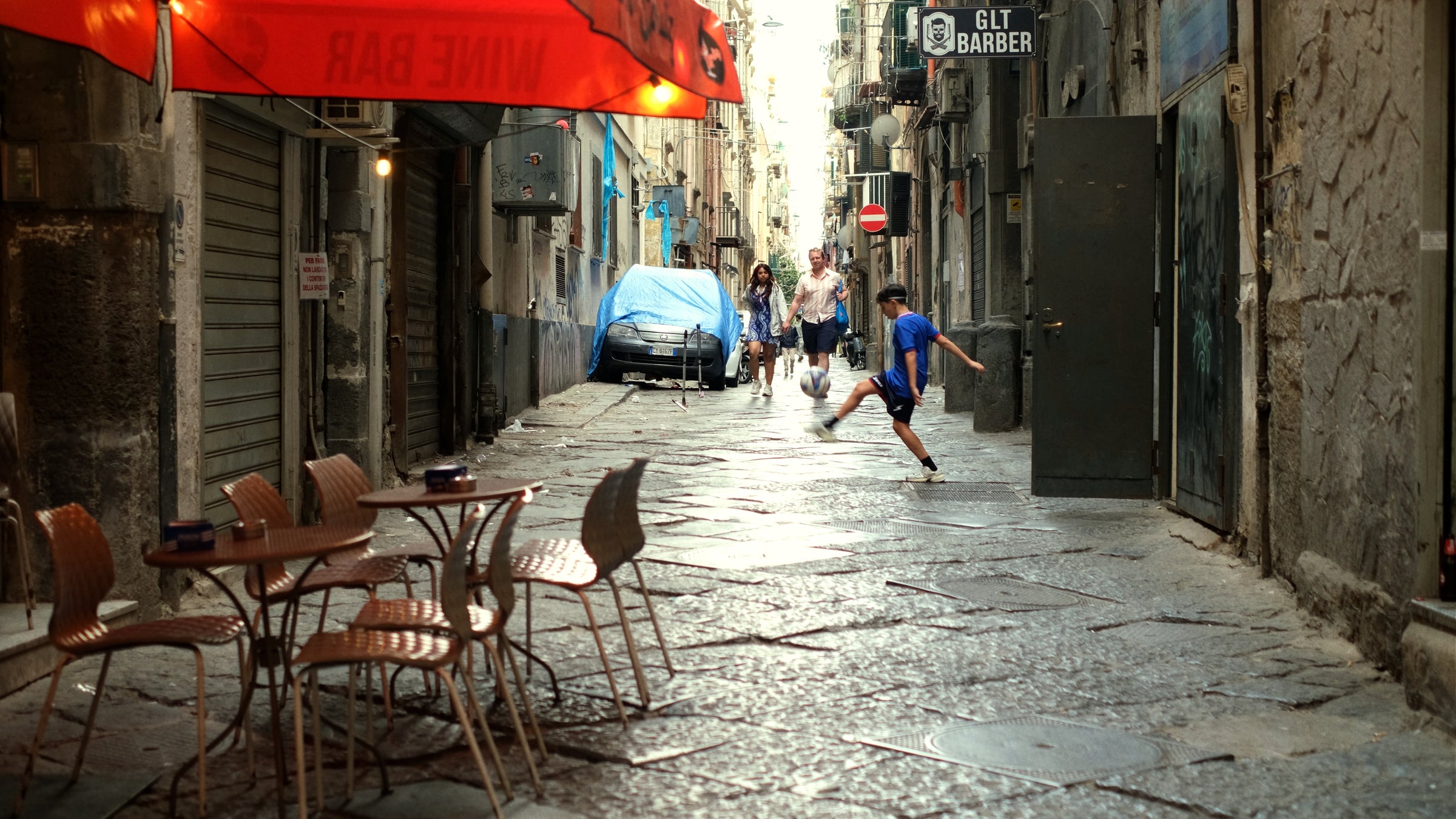 A narrow cobblestone alleyway with empty café tables in the foreground, a covered car in the background, and a boy in blue kicking a ball while people walk in the distance.