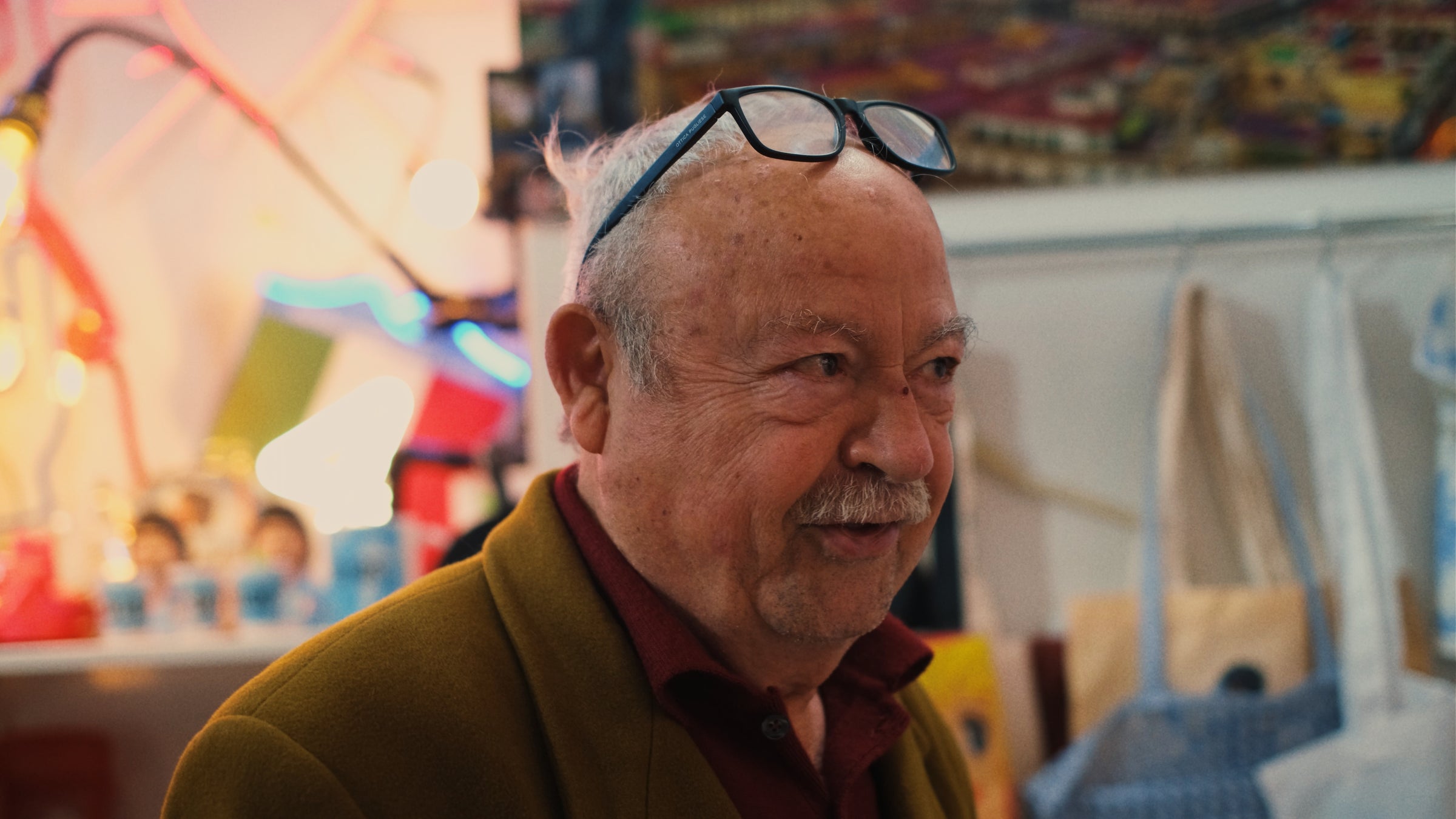 An elderly man with a mustache and glasses resting on his head smiles indoors, wearing a red shirt and brown jacket. Colorful objects and illuminated decorations are visible in the blurred background.