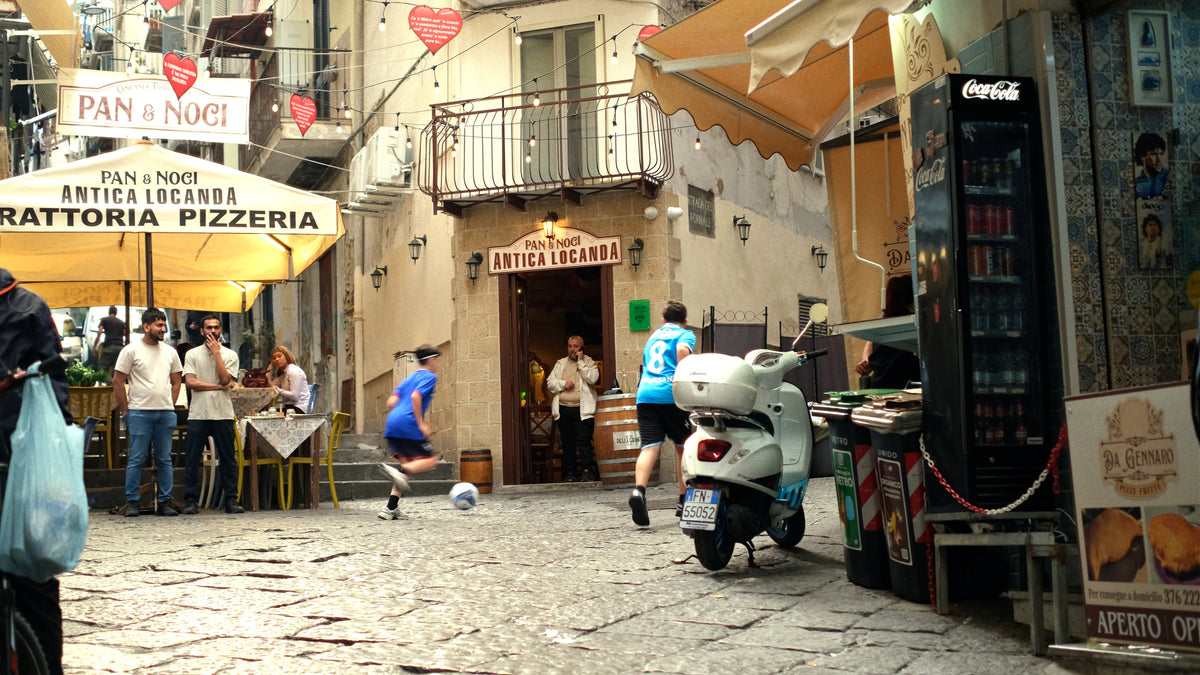 Two boys play soccer on a cobblestone street in front of a pizzeria, while people dine under yellow canopies and pedestrians stroll by in a lively, European outdoor setting.