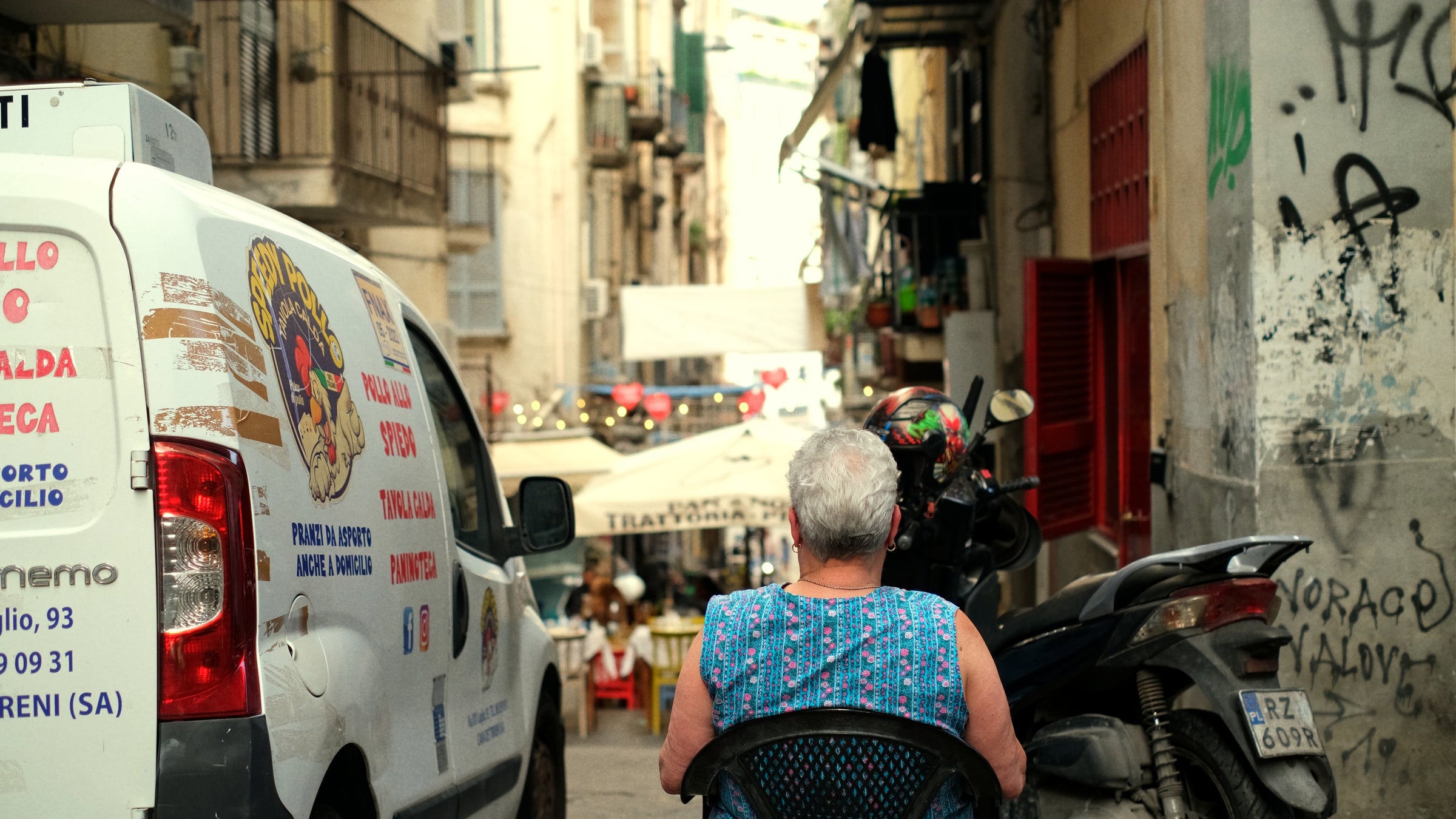 An elderly woman with short gray hair sits in a chair facing away on a narrow street lined with buildings, a van, motorcycles, and outdoor tables under umbrellas in the distance.