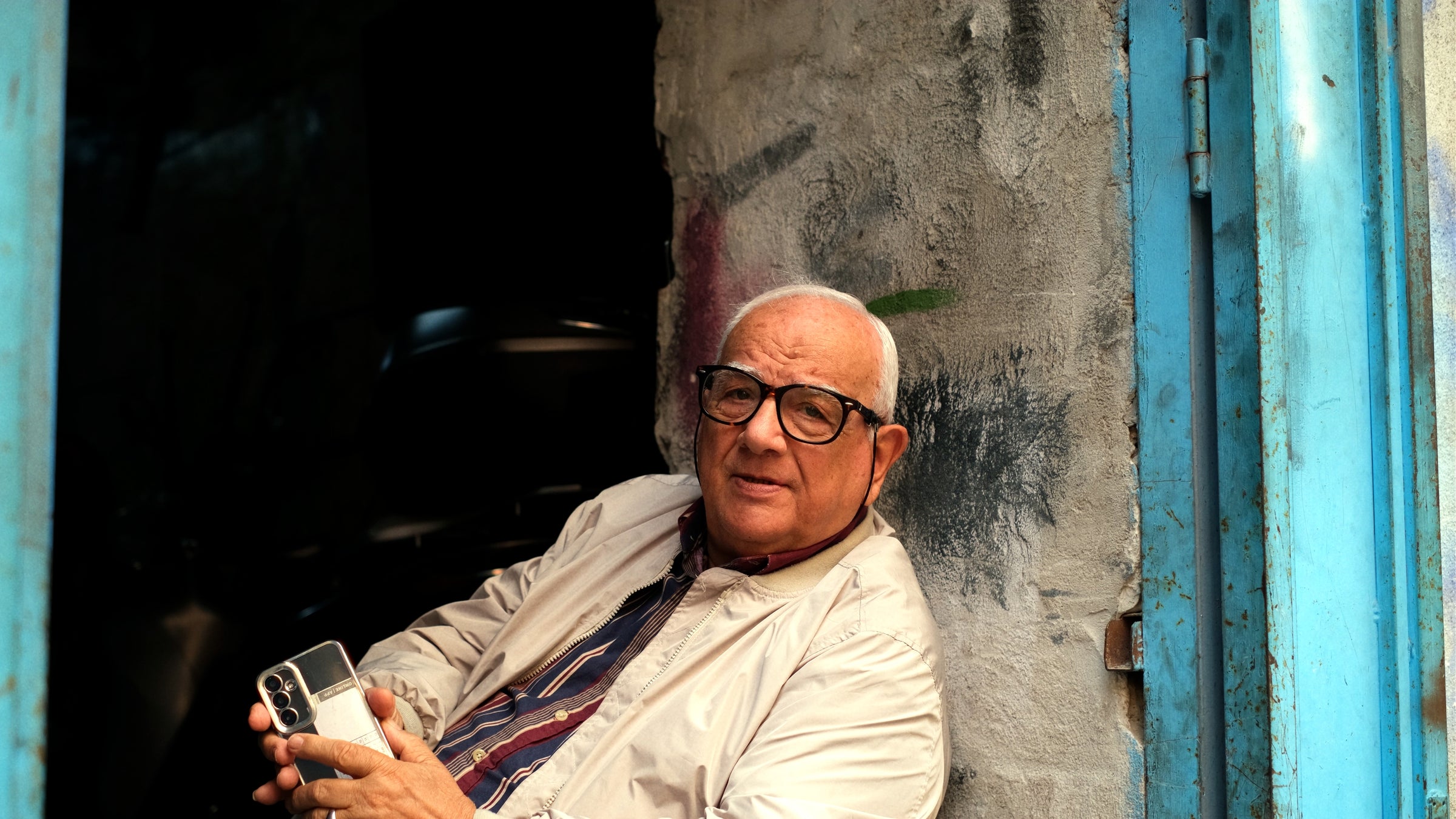 An older man with glasses and a white jacket sits in a doorway, holding a smartphone and looking at the camera. The doorframe is blue and the wall behind him is worn and textured.