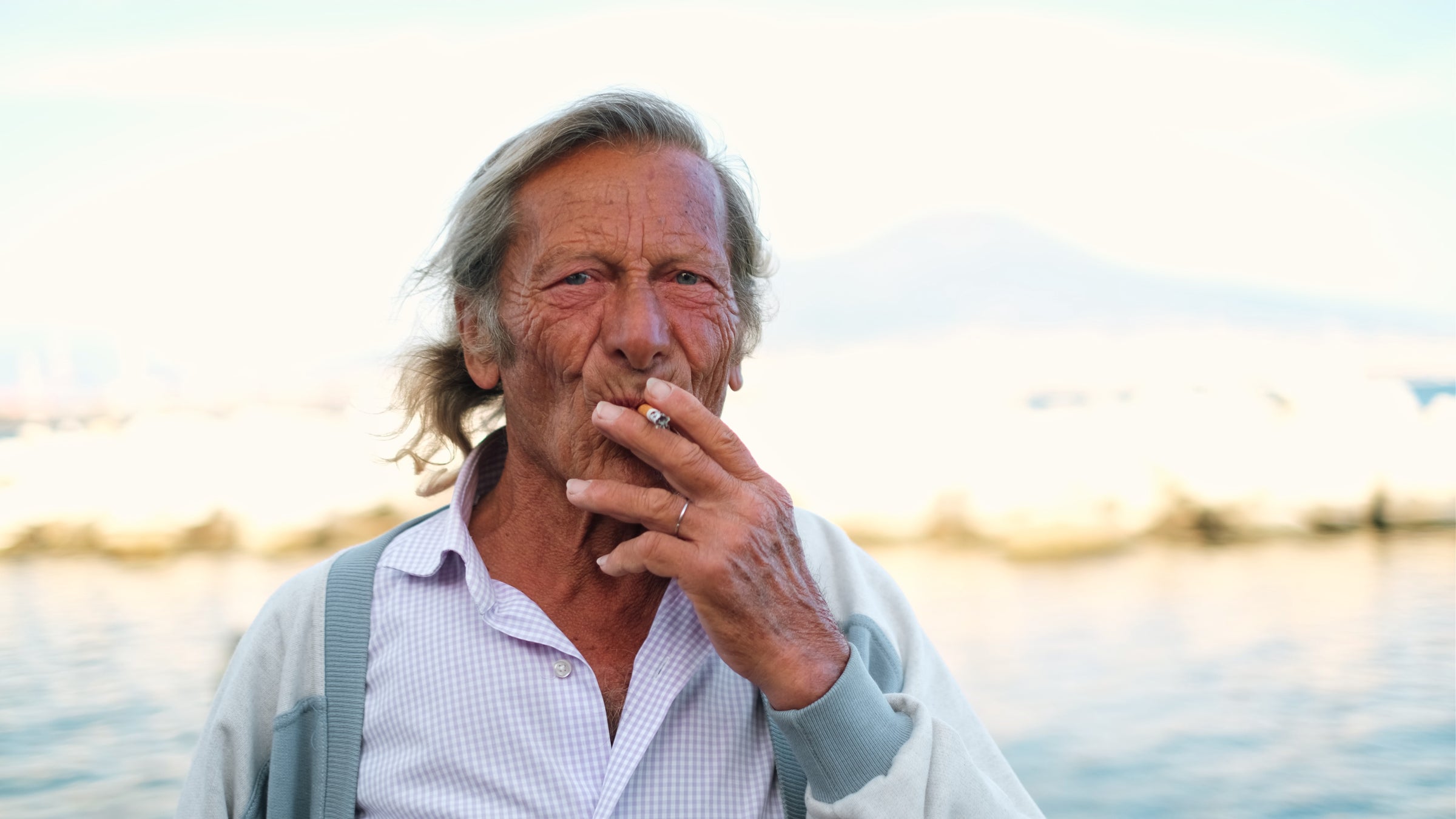 An older man with long gray hair is standing outdoors by the water, holding a cigarette to his mouth with one hand. He wears a light shirt and cardigan, with sunlight illuminating his face and a mountain blurred in the background.