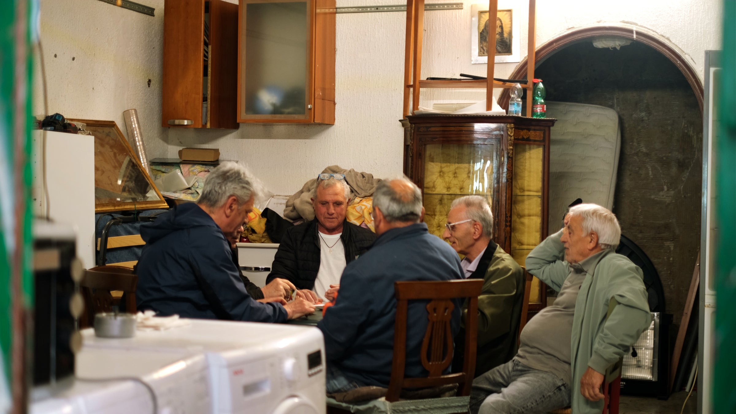 Five older men are sitting around a table in a cluttered, cozy room, engaged in conversation or a card game. The room has wooden cabinets, shelves, household items, and warm lighting.