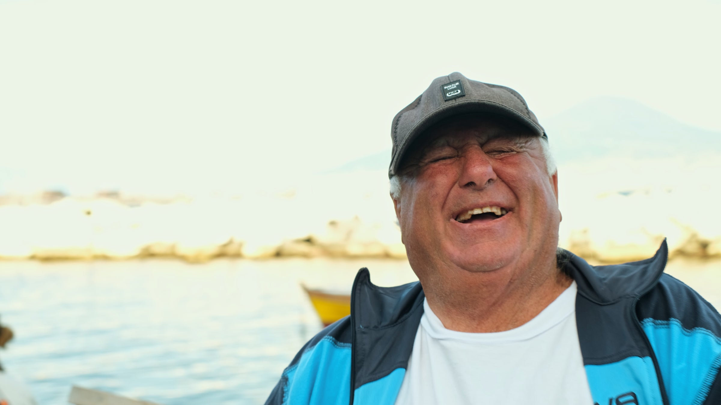 An older man wearing a baseball cap and blue jacket laughs joyfully outdoors, with a blurred waterfront and boat in the sunny background.