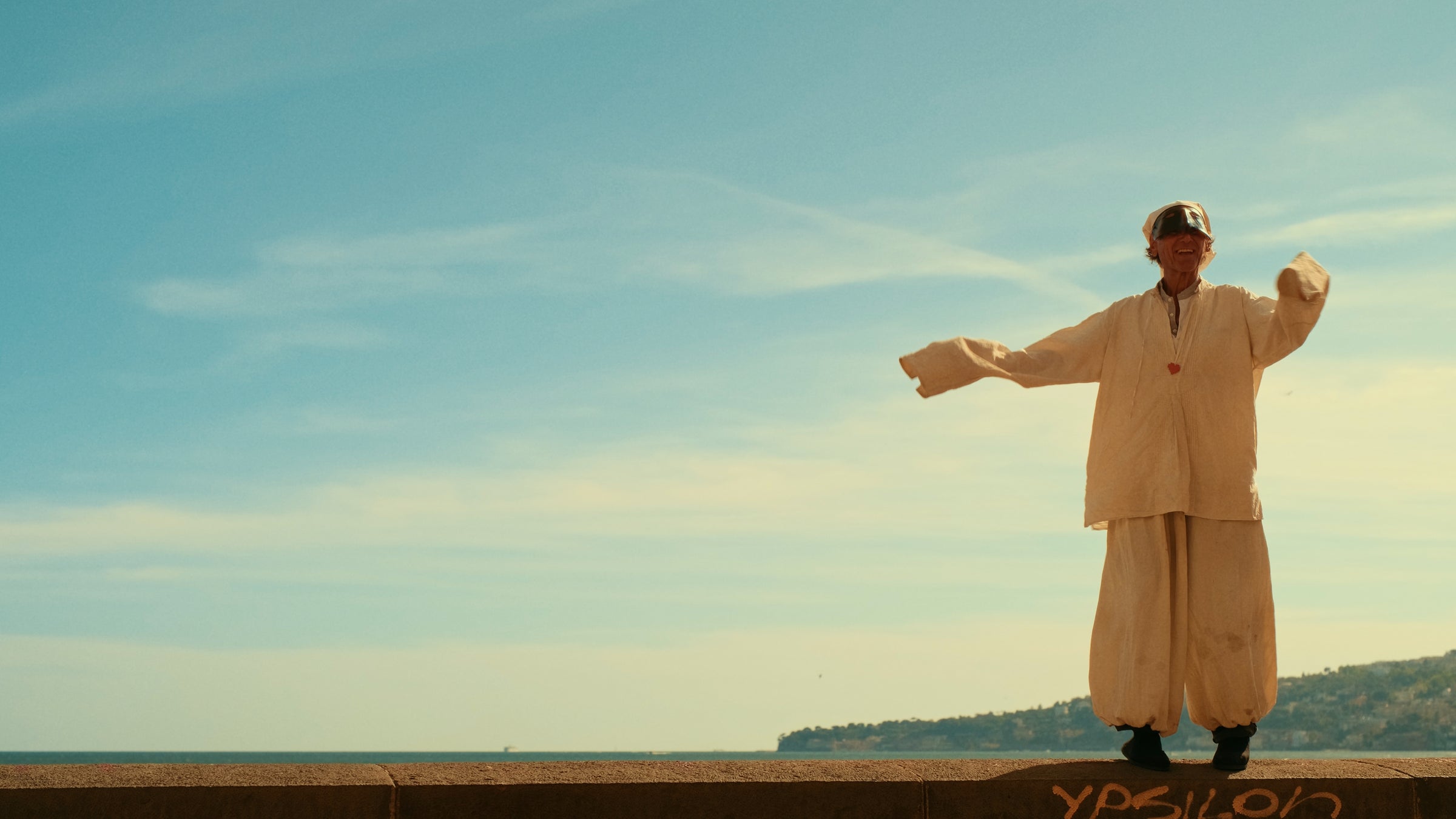 A person in loose, light-colored clothing and a hat stands on a ledge with arms outstretched against a blue sky, with distant hills and sea in the background.