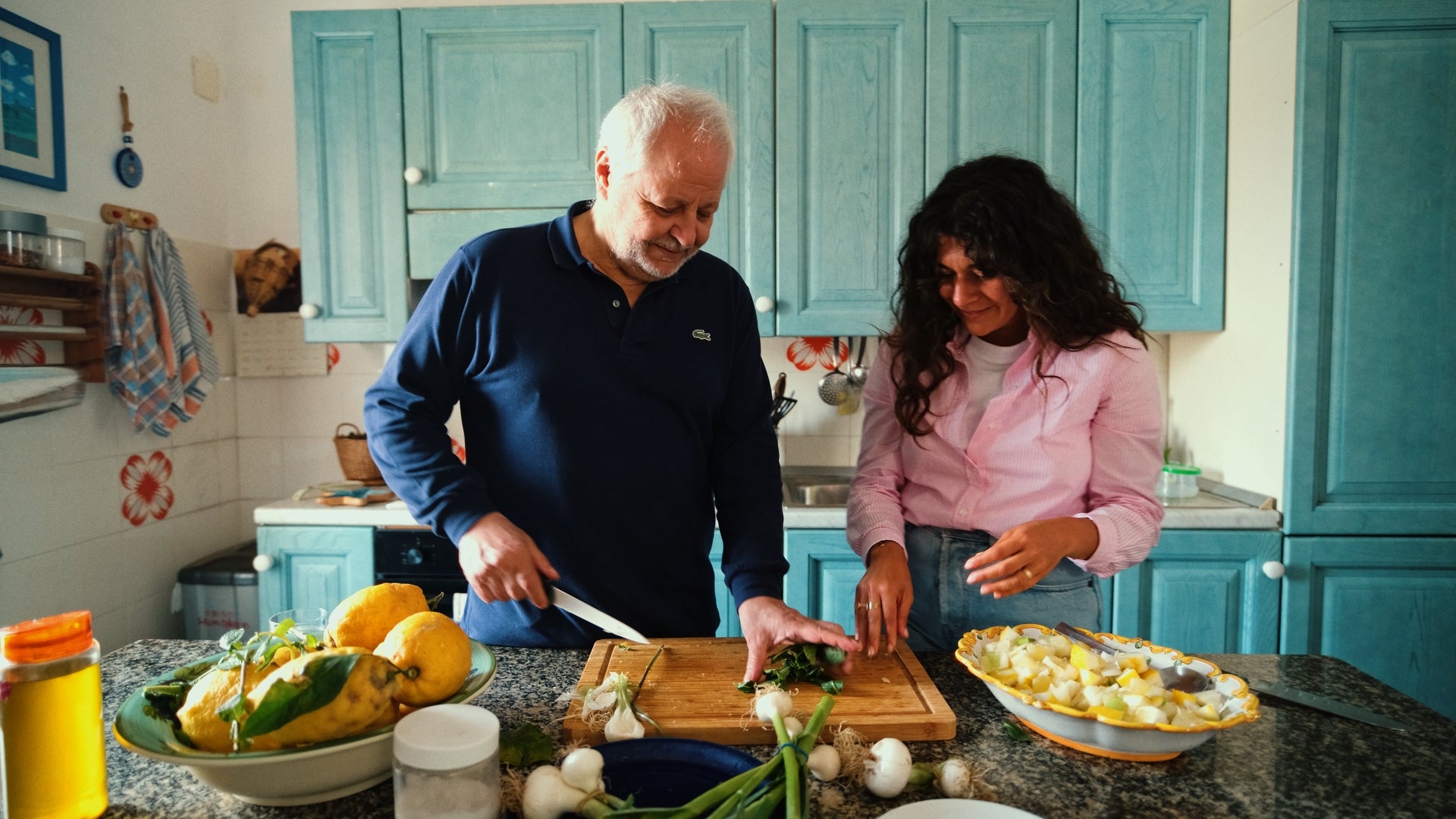 A man and a woman smile while preparing food together in a cozy kitchen with blue cabinets. The man chops herbs on a cutting board, while the woman stands nearby with chopped vegetables. Fresh produce is spread out on the counter.