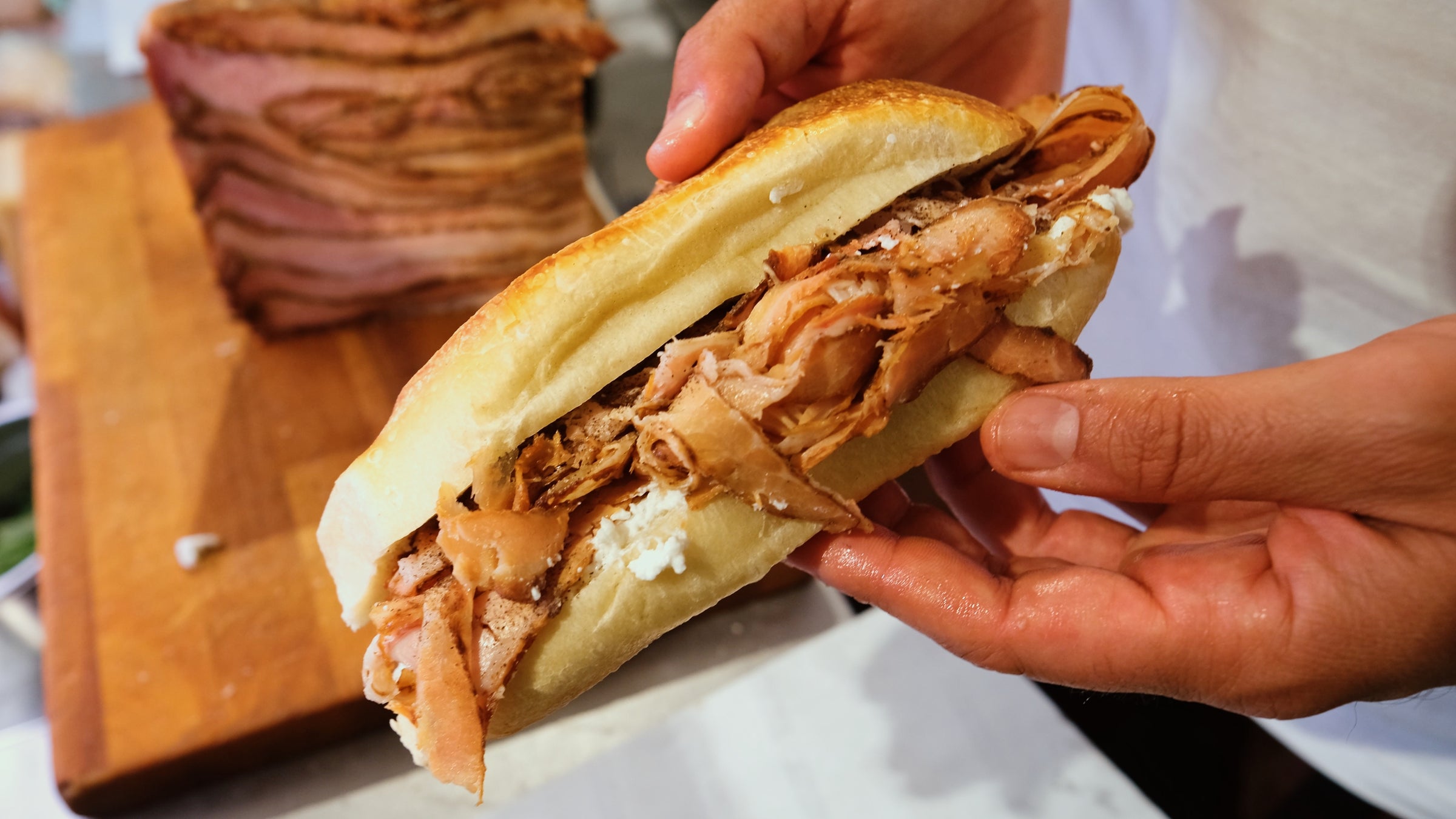 A person holds a sandwich filled with sliced roast meat and cheese on a crusty roll; more sliced meat is visible on a wooden cutting board in the background.