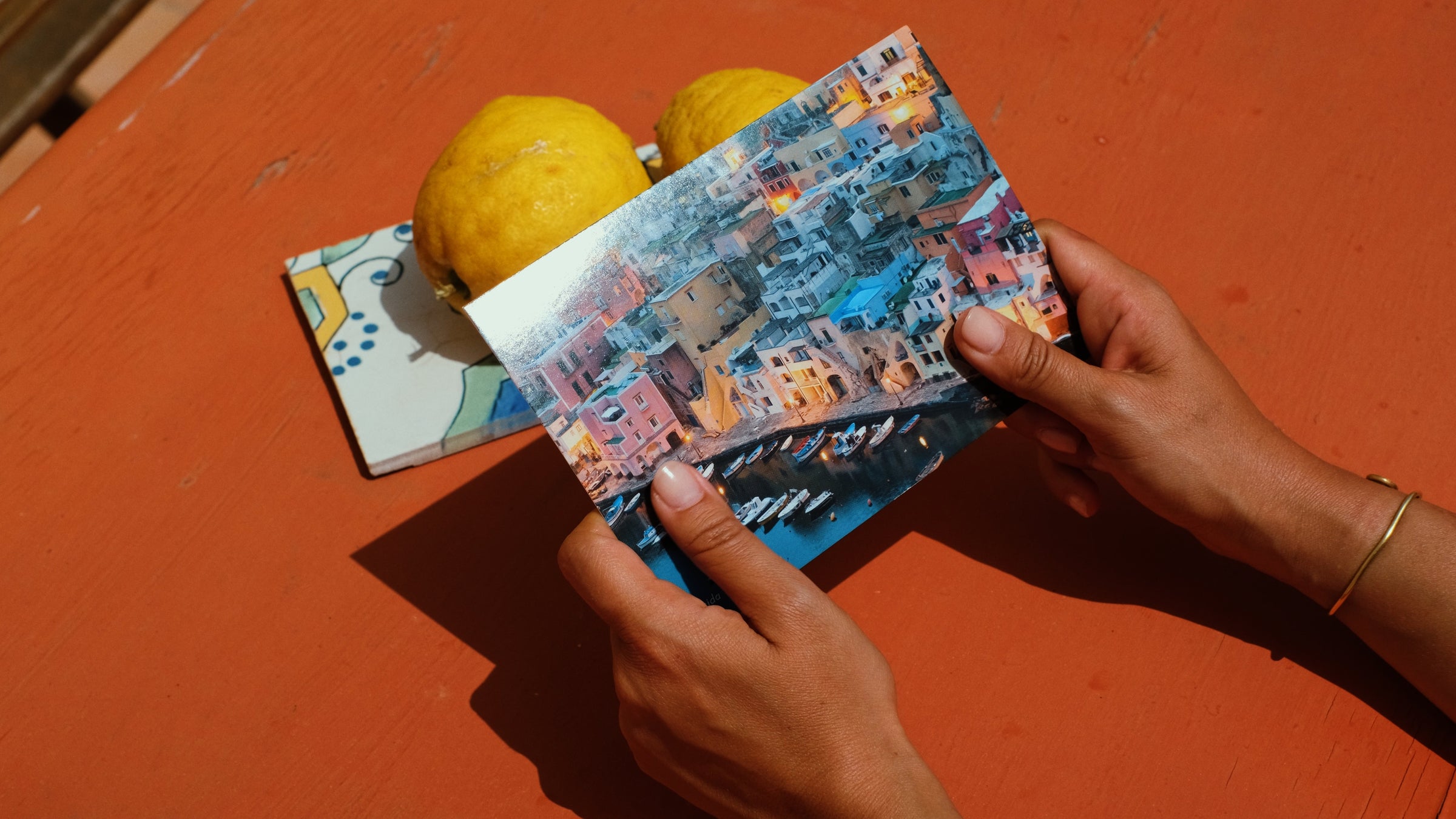 A person holds a postcard showing colorful seaside buildings. On the orange table are two lemons and a decorative tile. The person wears a thin bracelet on one wrist.