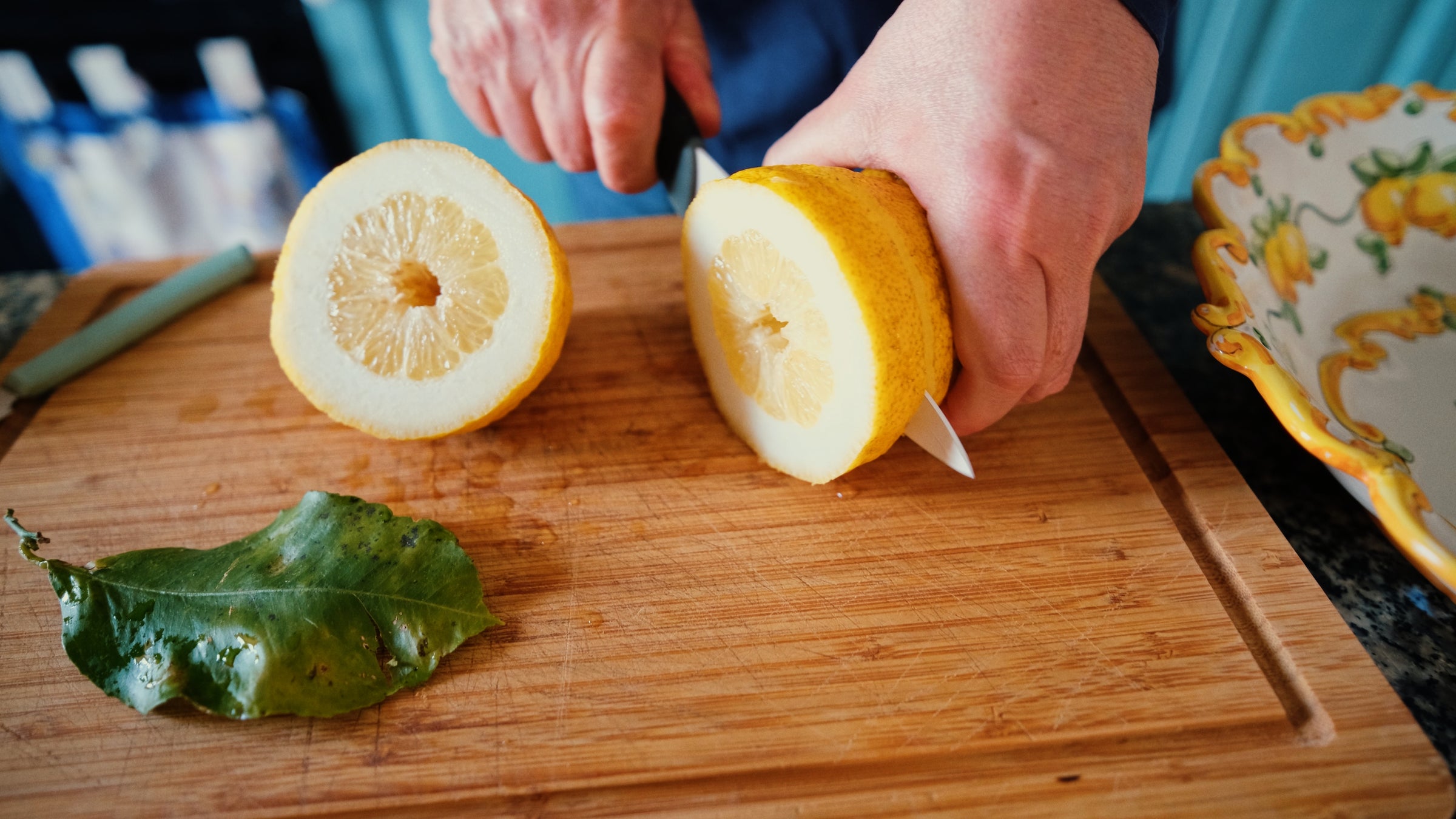 A person slices a large yellow lemon in half on a wooden cutting board, with a green lemon leaf and a decorative dish nearby.