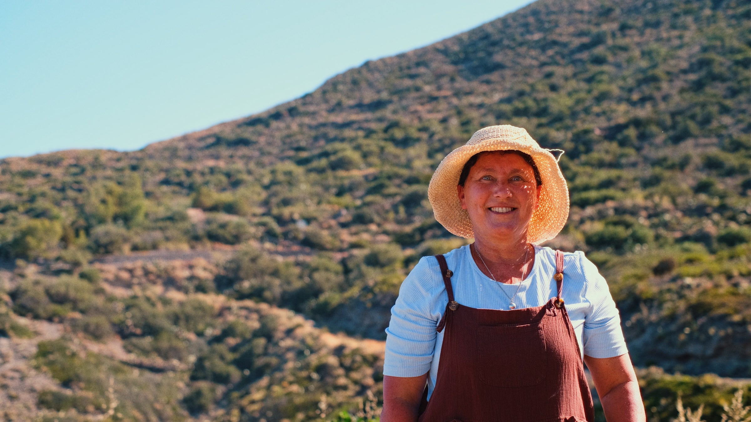 Smiling woman in a straw hat and overalls stands outdoors with green hills and blue sky in the background.