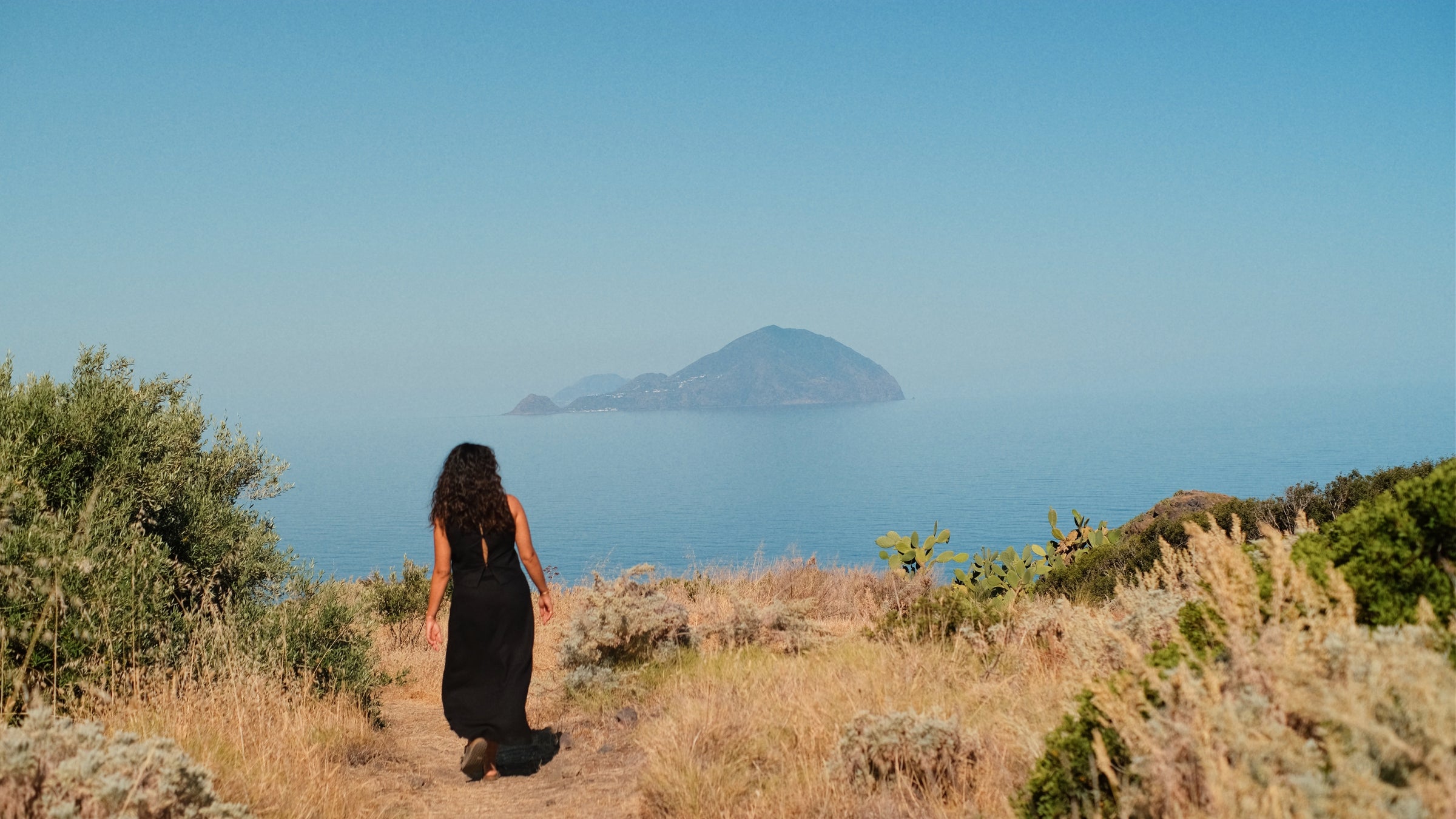 A woman in a black dress walks along a path through dry, grassy terrain toward the ocean, with an island visible in the distance under a clear blue sky.
