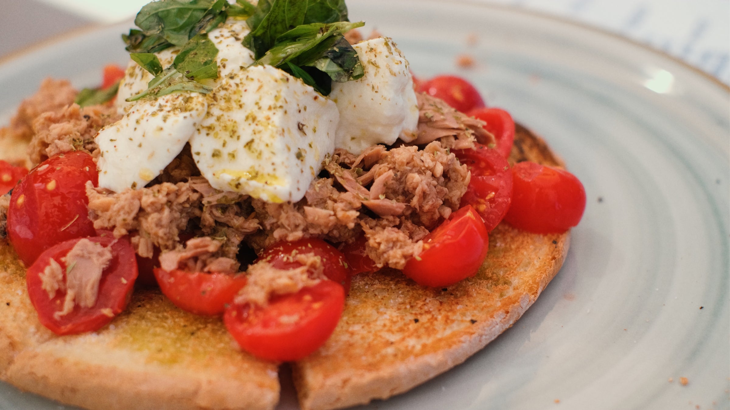 Toasted bread topped with cherry tomatoes, flaked tuna, mozzarella cheese, fresh herbs, and a sprinkle of seasoning, served on a light-colored plate.