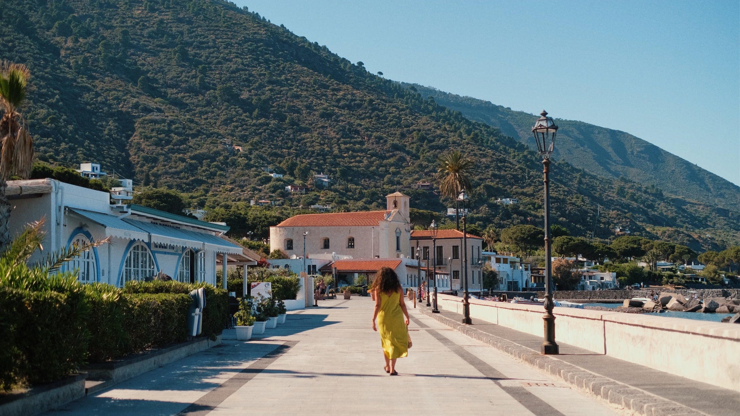 A person in a yellow dress walks along a sunny coastal promenade lined with buildings, with a church and lush green mountains in the background.