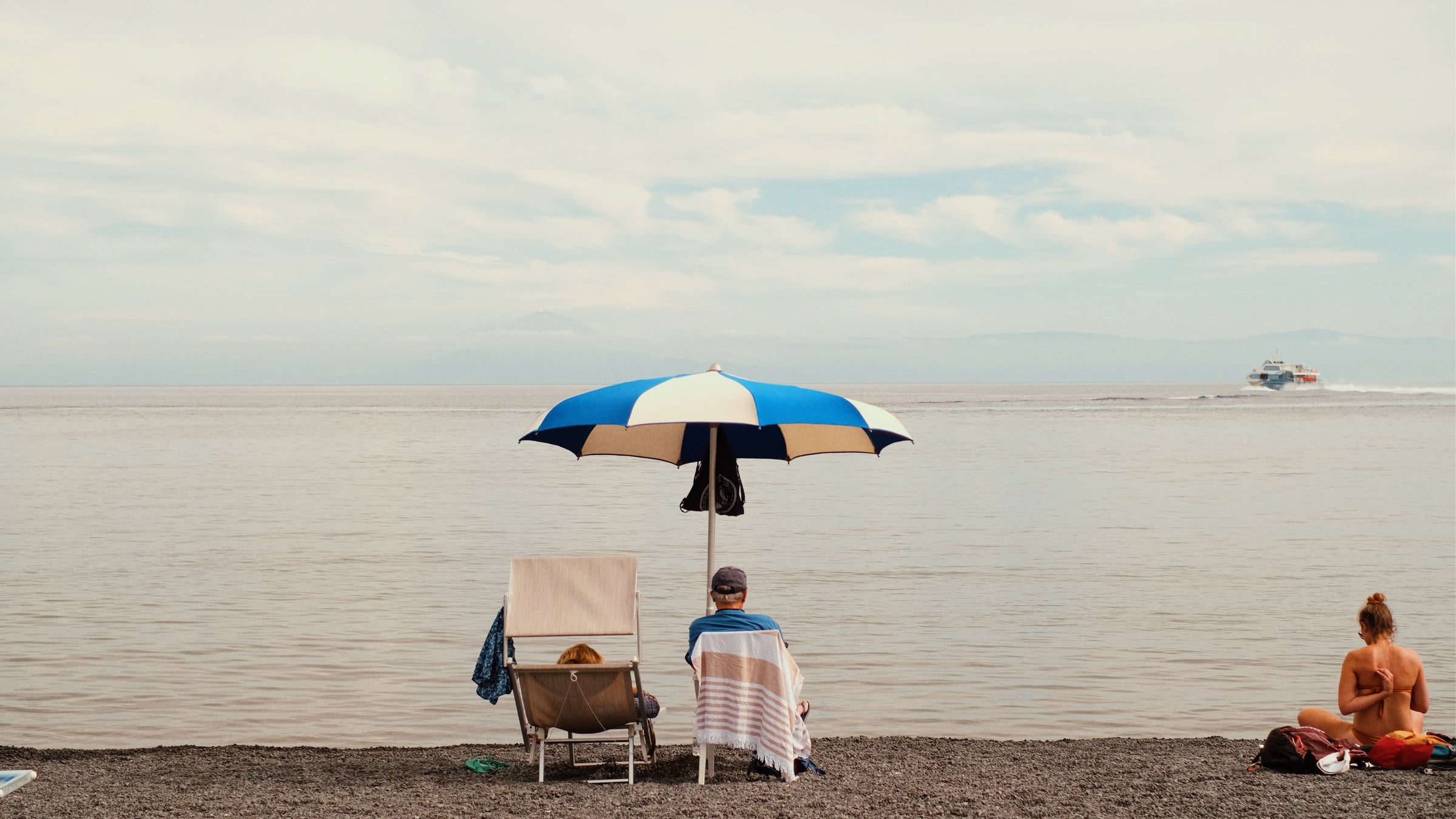 A person sits under a blue and white umbrella on a beach facing the water, with a towel draped over their chair. Another person sits on the right, and a boat is visible in the distance.