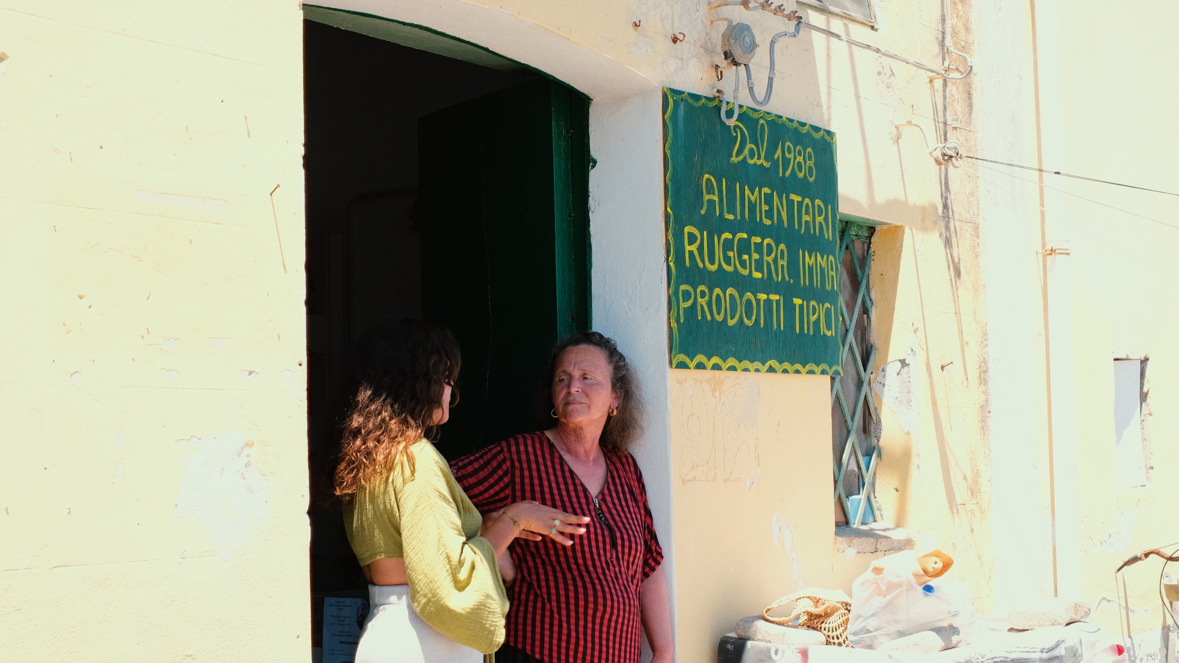 Two people stand and talk in the doorway of a light yellow shop with a green sign that reads Dal 1988 Alimentari Ruggeri Imma Prodotti Tipici. Sunlight shines on the scene.