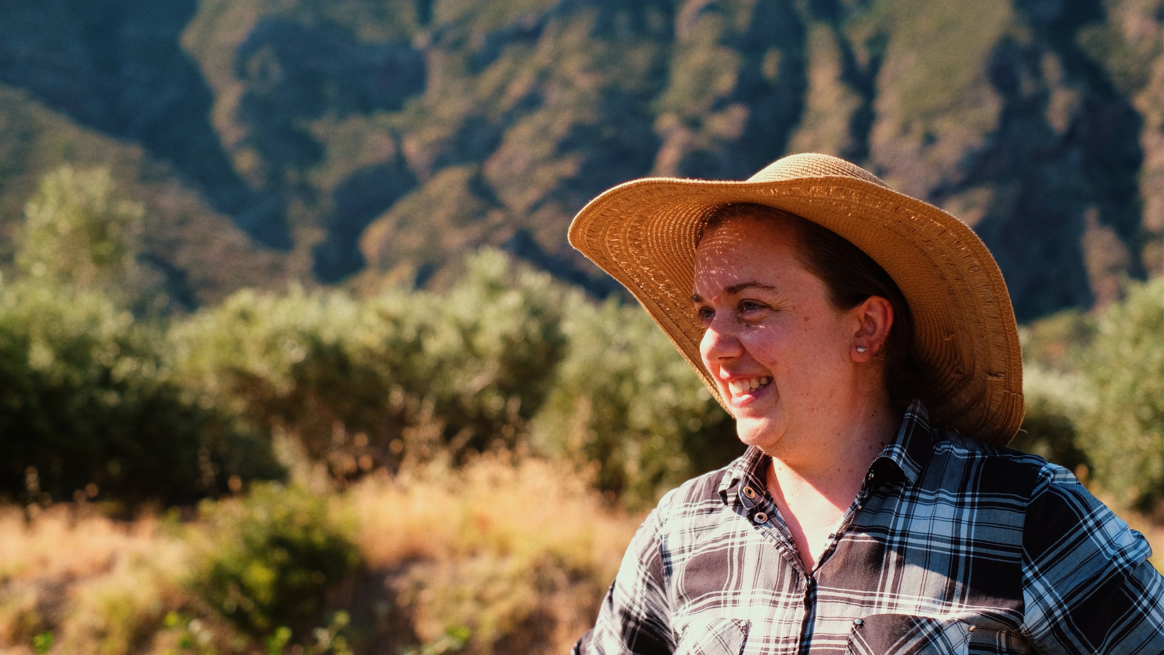 A woman wearing a straw hat and plaid shirt smiles outdoors, with sunlit mountains and greenery in the background.