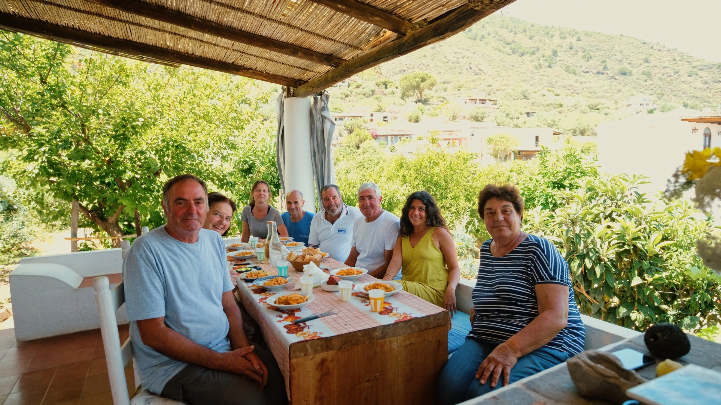 A group of nine people sit around a table outdoors under a shaded patio, sharing a meal. Trees and hills are visible in the background on a sunny day.