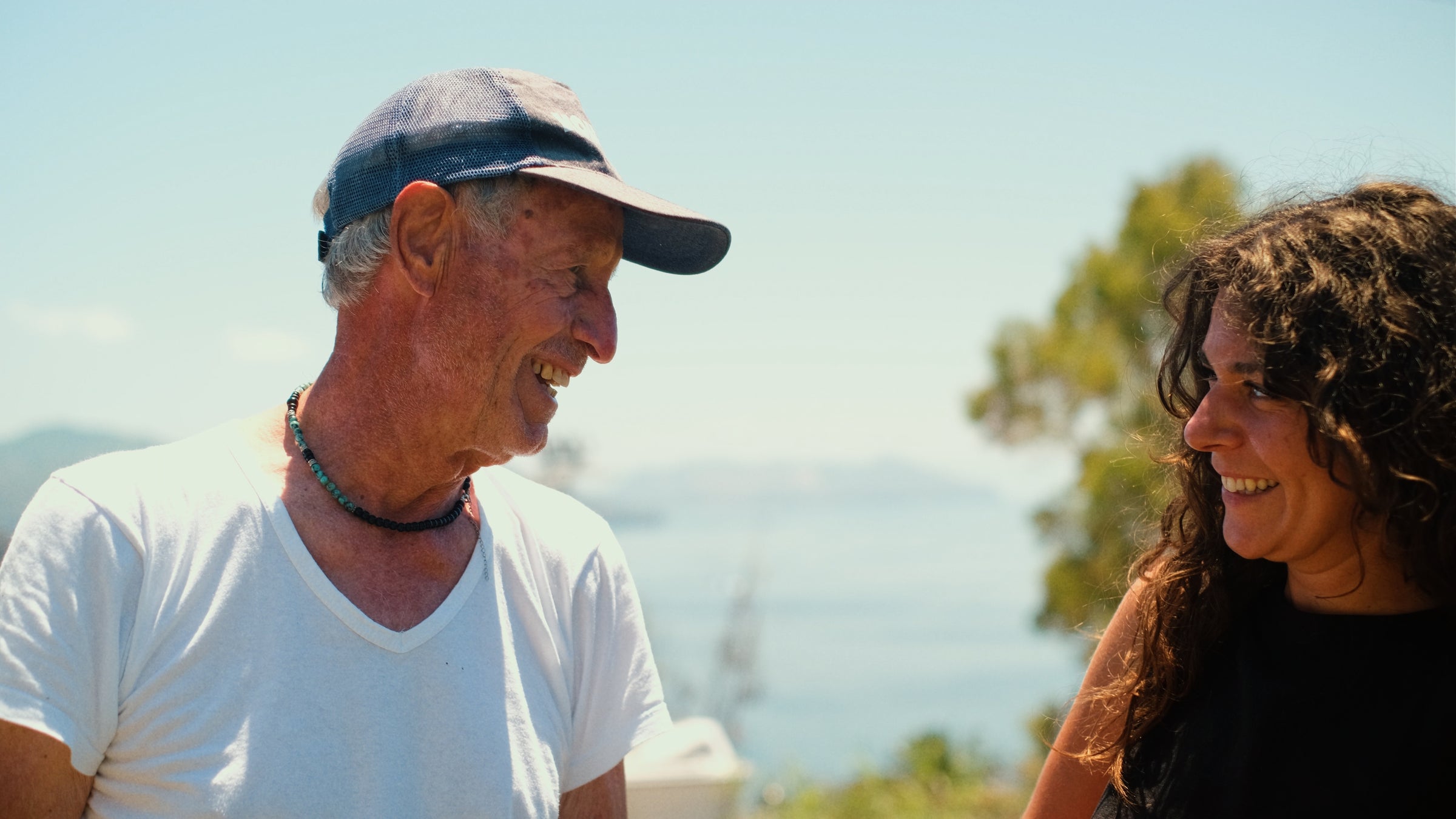 An older man in a white shirt and cap smiles at a woman with long curly hair. They stand outdoors with trees and a blurred view of water and mountains in the background, both appearing happy and joyful.