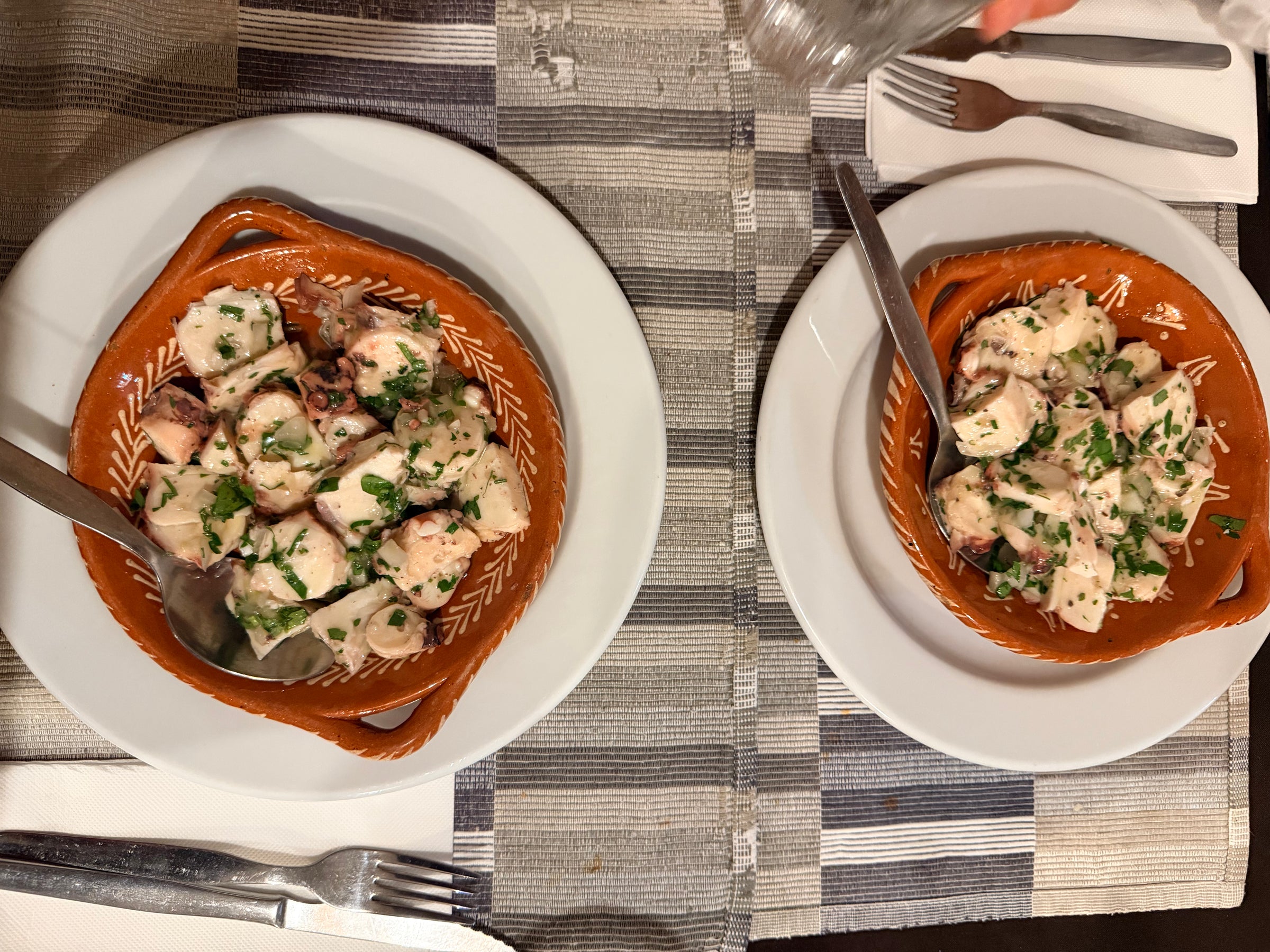 Two terracotta dishes on white plates, each filled with octopus pieces garnished with chopped parsley. Spoons rest in the dishes, and a hand is sprinkling salt on one dish. Forks and napkins are placed beside the plates.
