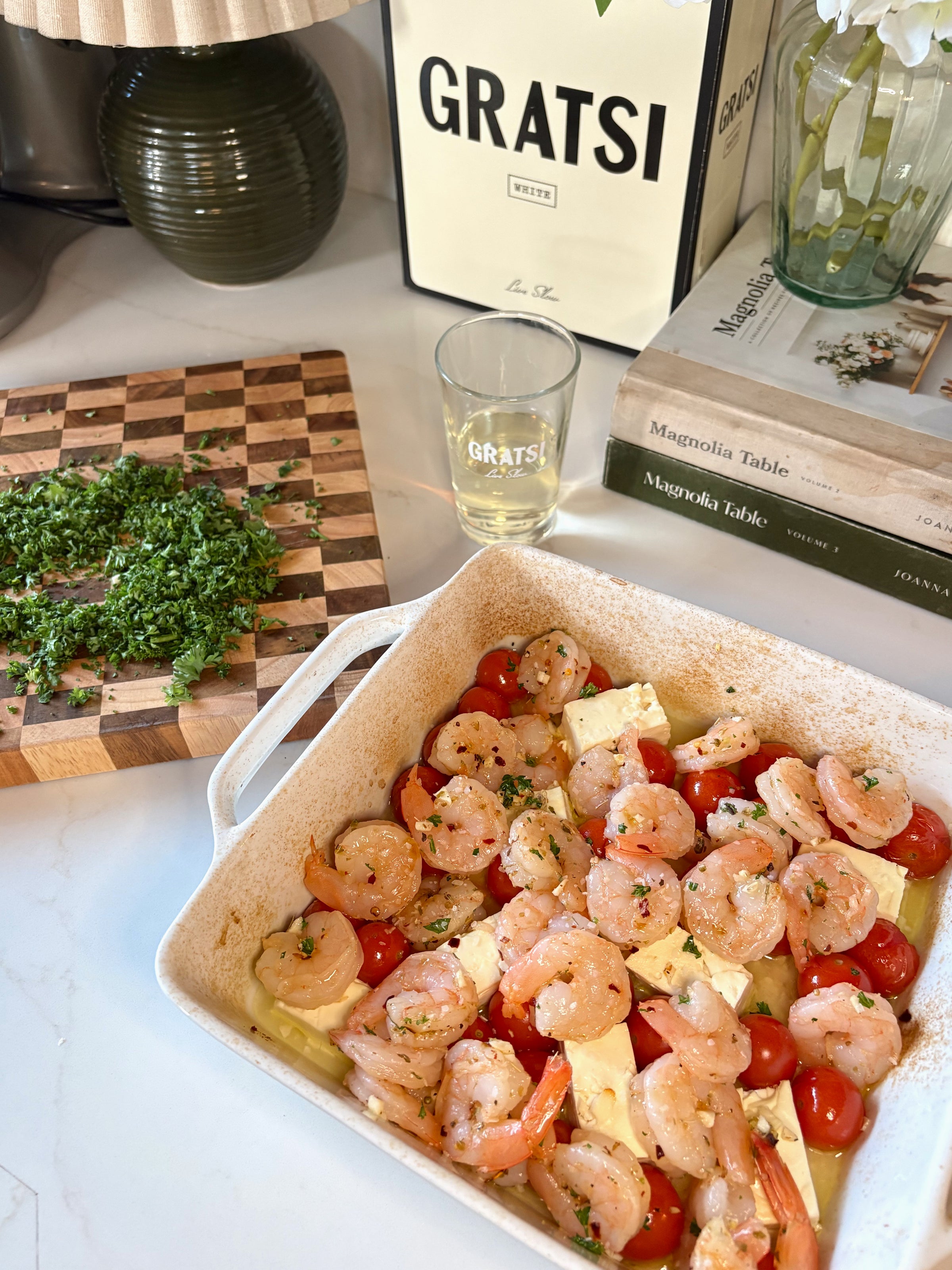 A baking dish filled with shrimp, cherry tomatoes, and feta cheese sits on a white counter. Nearby are chopped herbs on a cutting board, a glass of white wine, books, and a Gratsi wine box.