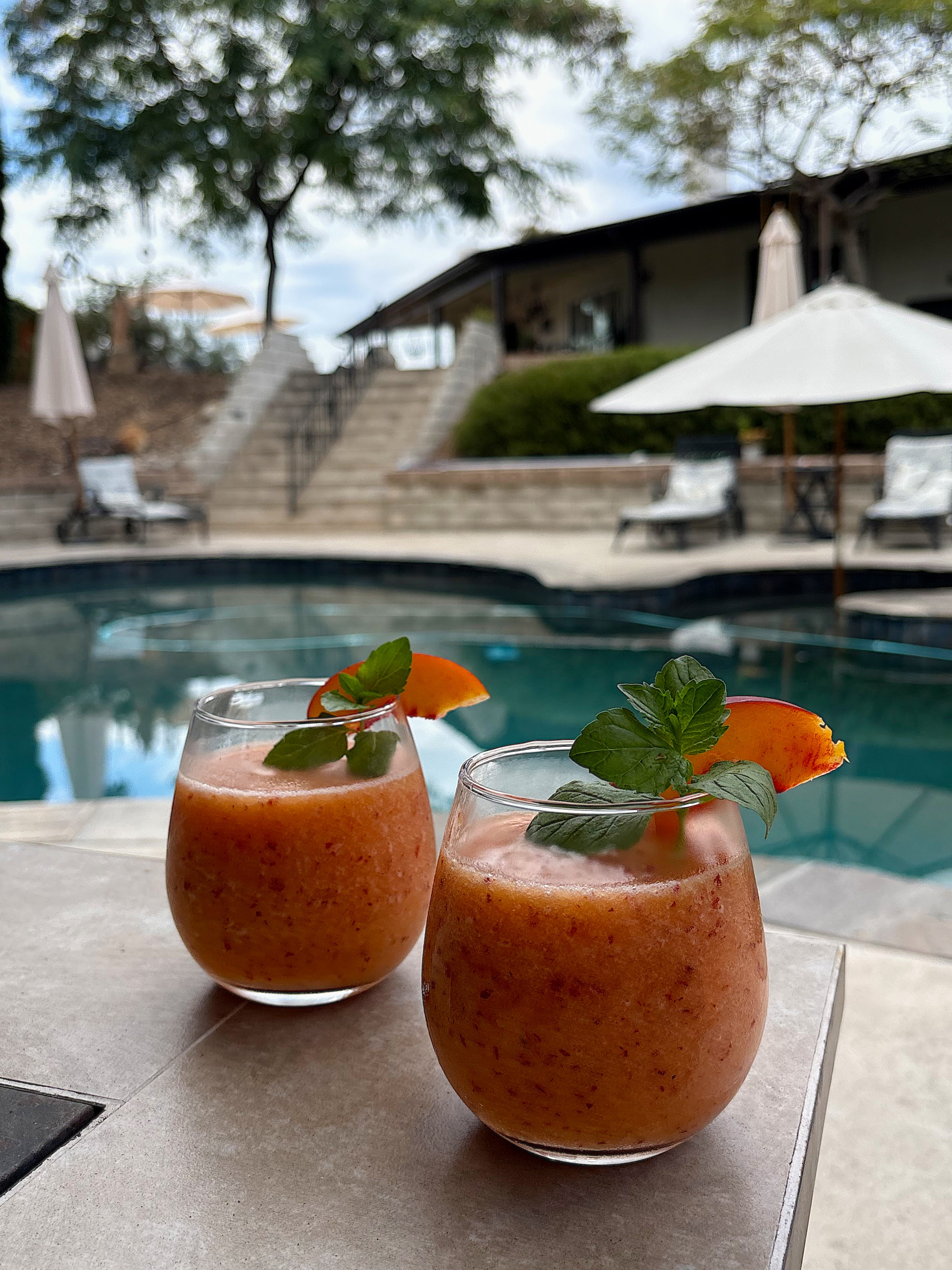 Two glasses of peach smoothie garnished with mint and peach slices sit on a table beside a pool, with lounge chairs, umbrellas, and trees in the background.