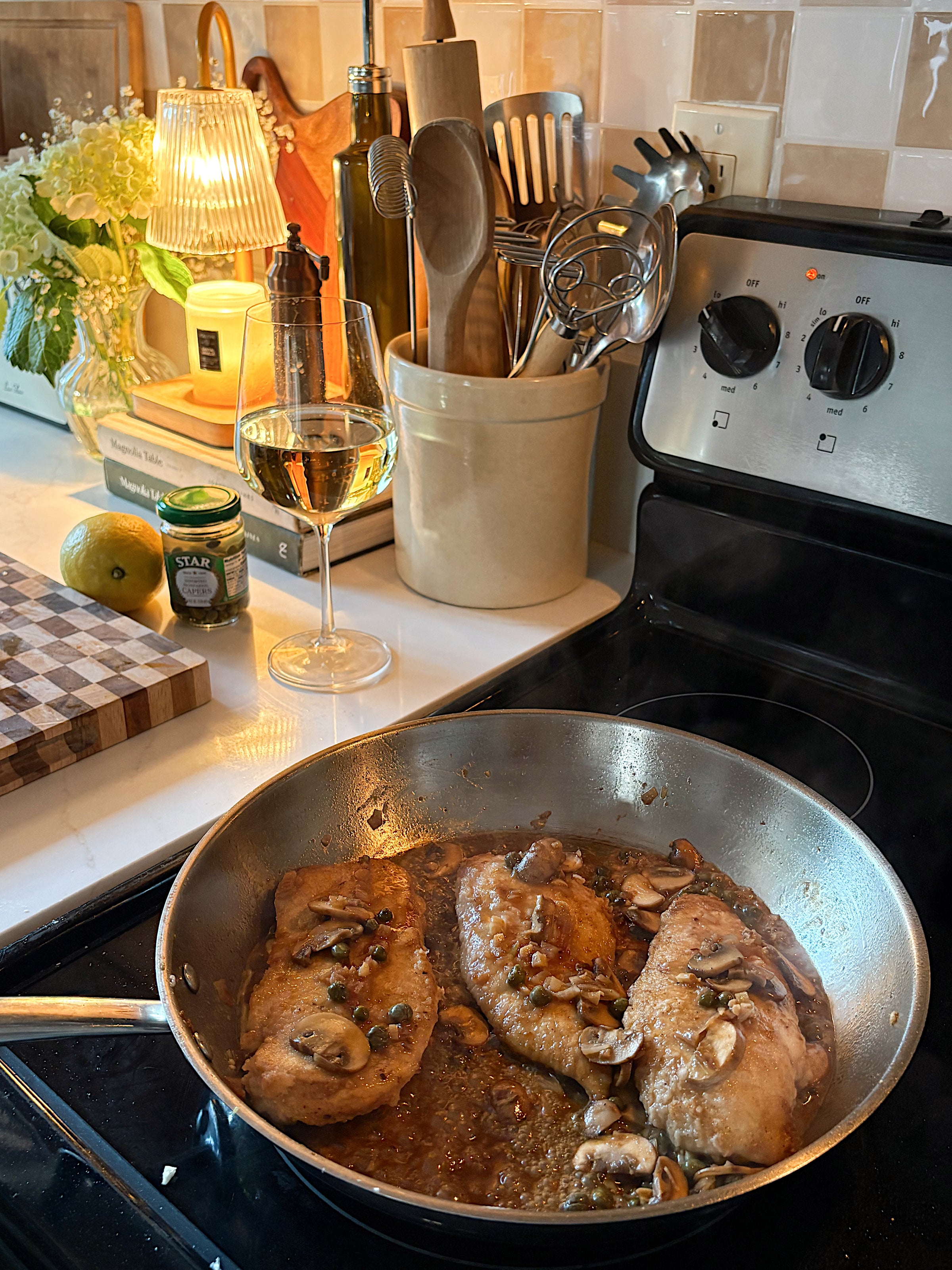 Chicken breasts with mushrooms cook in a sauté pan on a stove. A glass of white wine, kitchen utensils, olive oil, and herbs are nearby, creating a cozy, homey kitchen scene.
