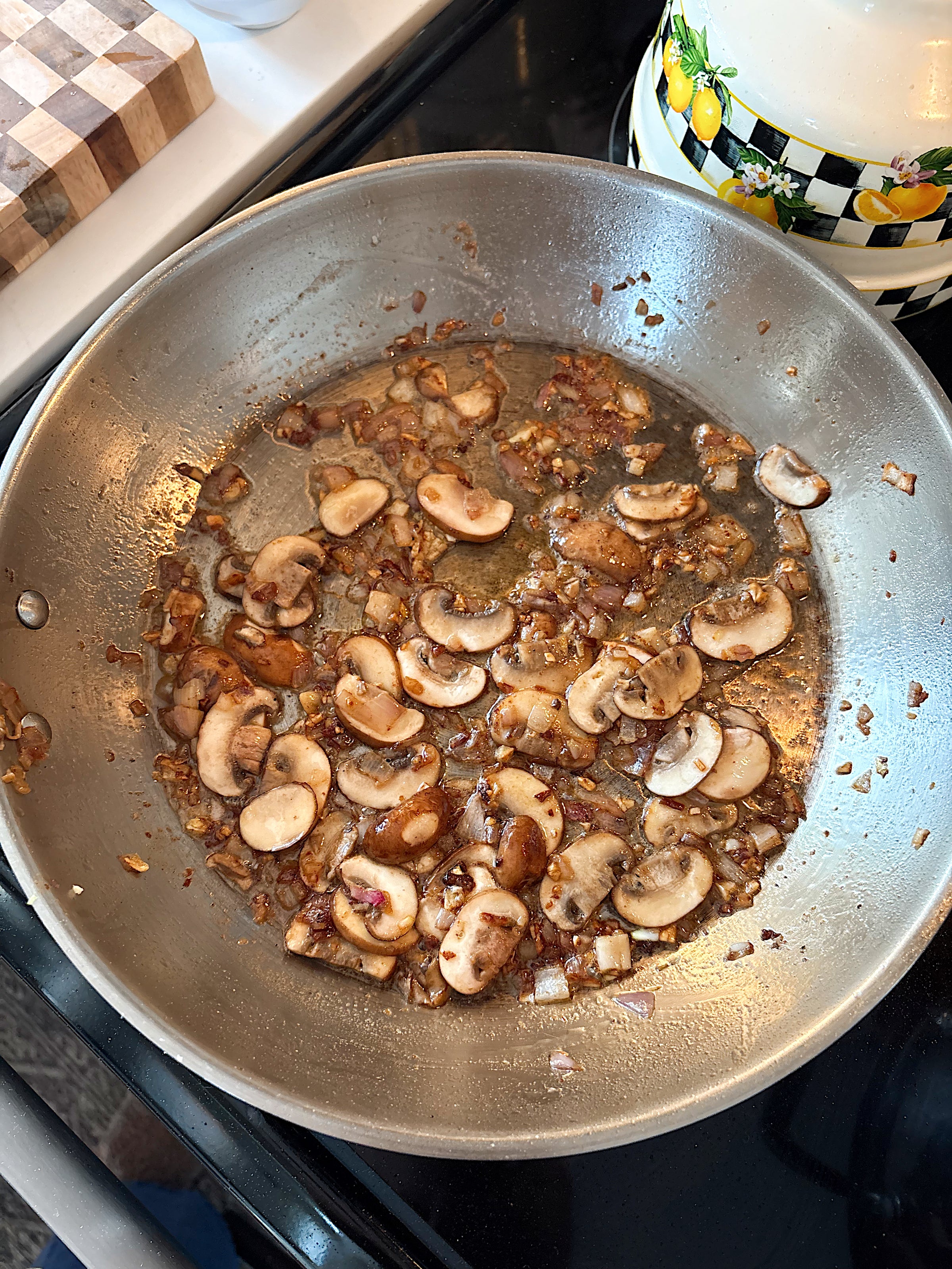 Sliced mushrooms and diced onions are being sautéed in a stainless steel pan on a stovetop, with some oil and seasonings visible. A patterned teapot and wooden cutting board are nearby.