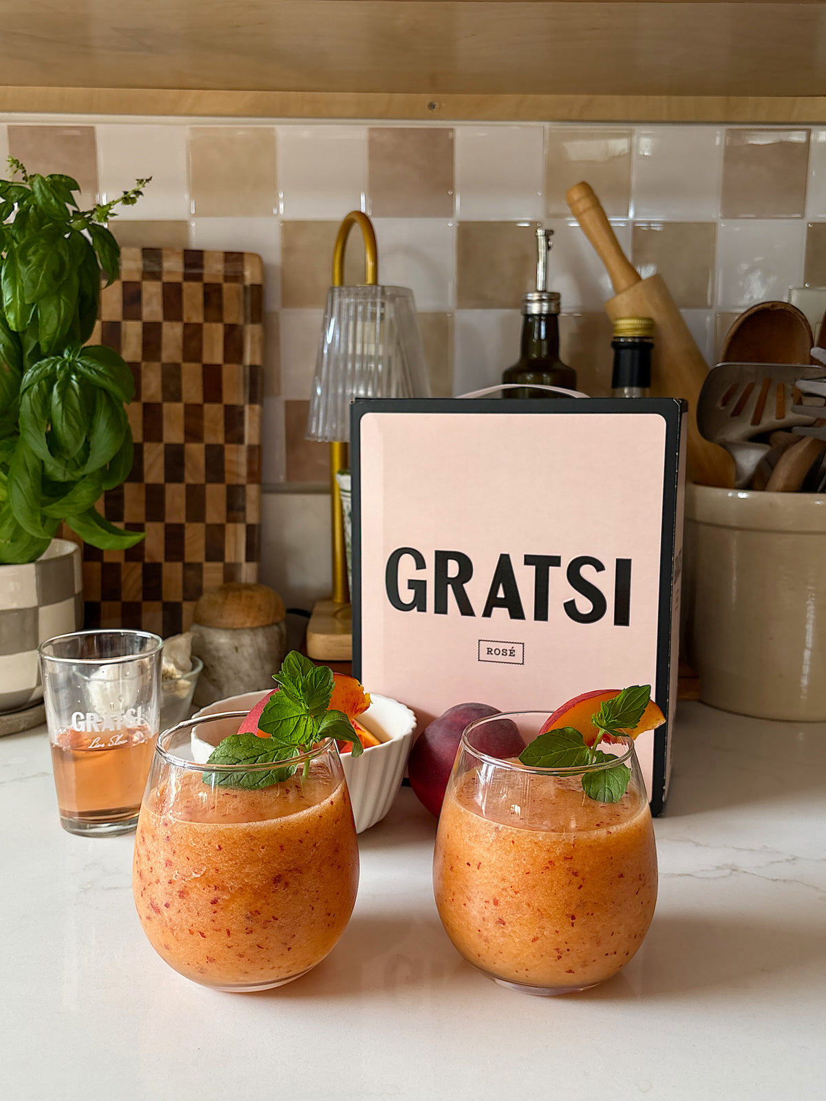 Two peachy-orange drinks with mint garnish sit on a kitchen counter. Behind them are peaches, a box labeled GRATSI, a small glass, a basil plant, and various kitchen utensils. The scene looks fresh and inviting.