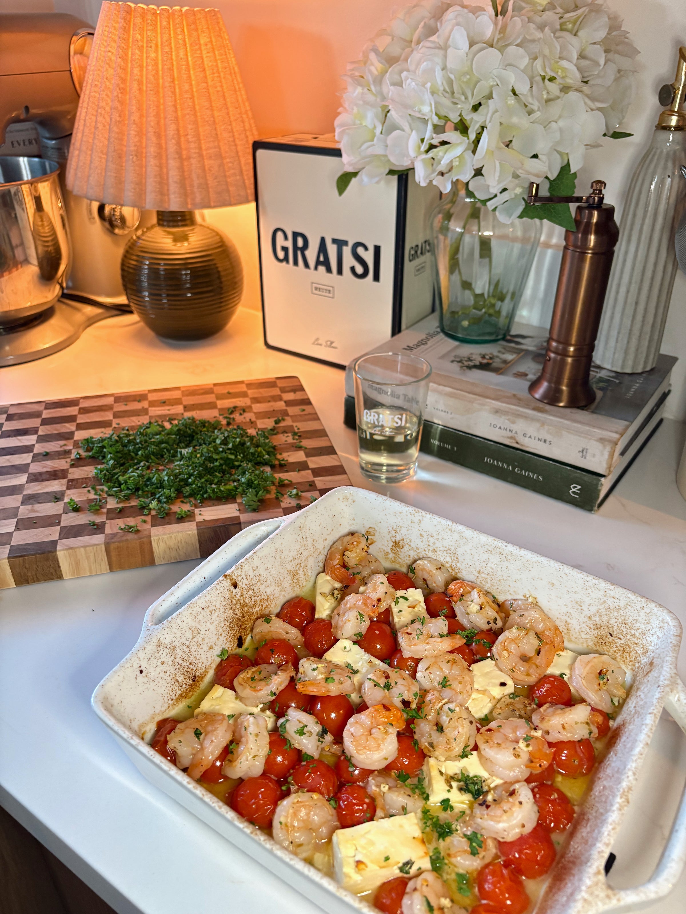 A dish of baked shrimp, cherry tomatoes, and feta cheese is garnished with herbs on a kitchen counter. Nearby are a cutting board with chopped herbs, a glass of water, cookbooks, a lamp, and a vase of white flowers.