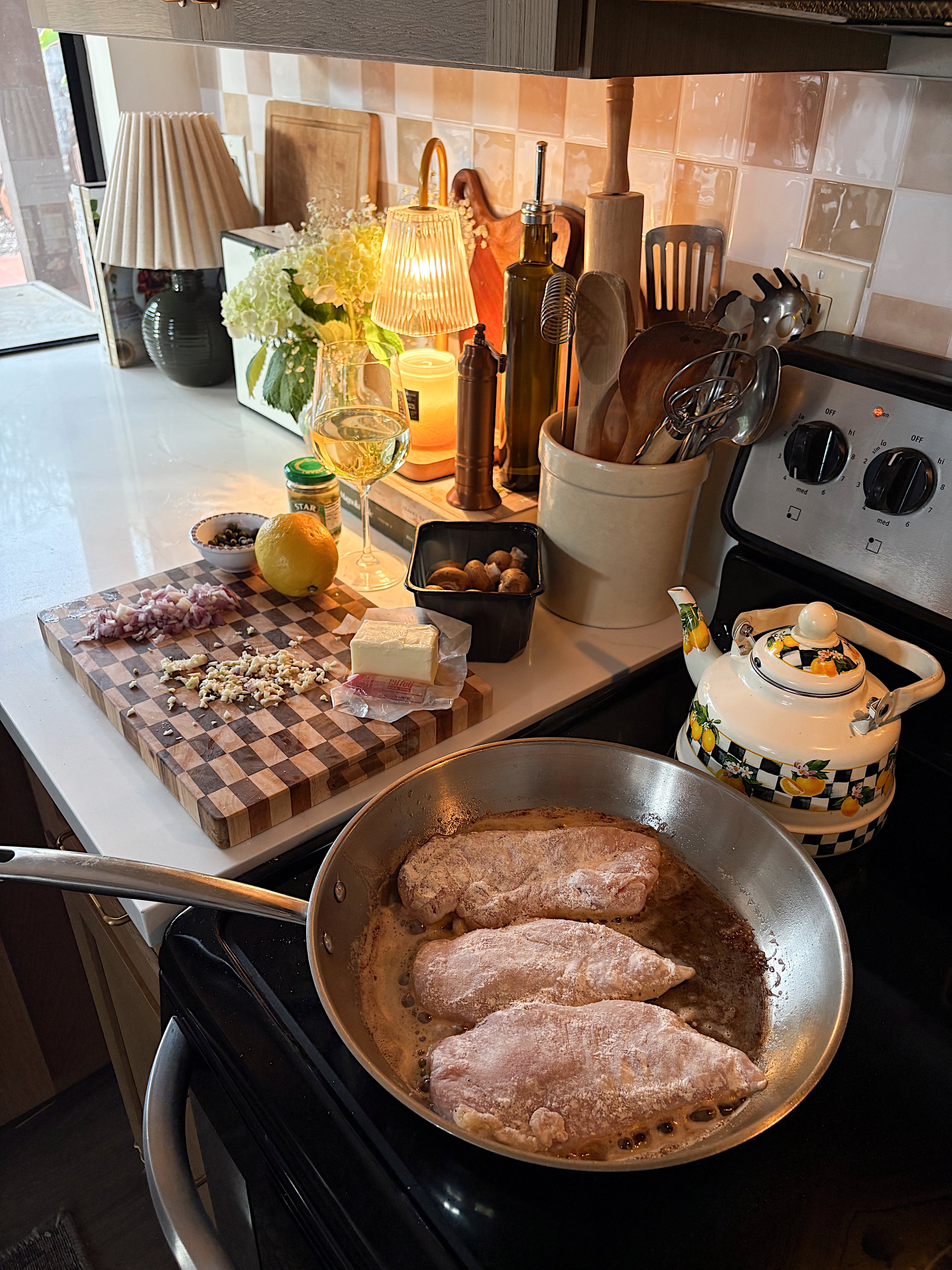Three breaded chicken breasts cook in a frying pan on a stove. Nearby, a teapot sits on another burner, and a cutting board holds chopped shallots, mushrooms, lemon, garlic, and cheese. Kitchen utensils and a wine glass are in the background.