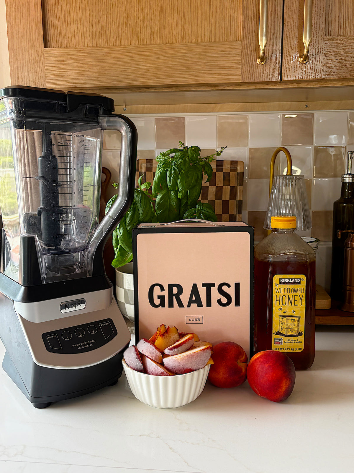 A blender, a bowl of sliced nectarines, whole nectarines, a jar of honey, basil, and a pink “GRATSI” cookbook are arranged on a kitchen counter with wood cabinets and a tiled backsplash.