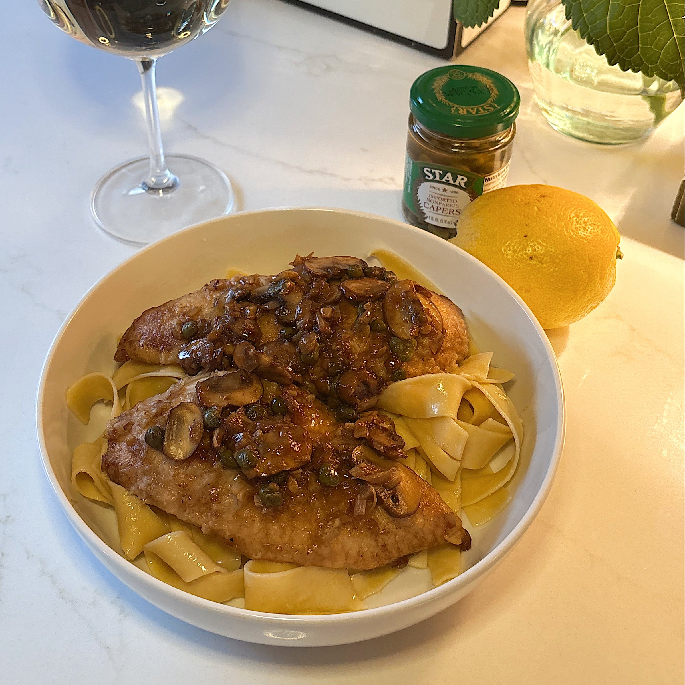 A bowl of pappardelle pasta topped with two pieces of chicken and sautéed mushrooms sits on a white counter next to a glass of white wine, a lemon, a small jar, and a vase of white flowers. A GRATSI sign is in the background.