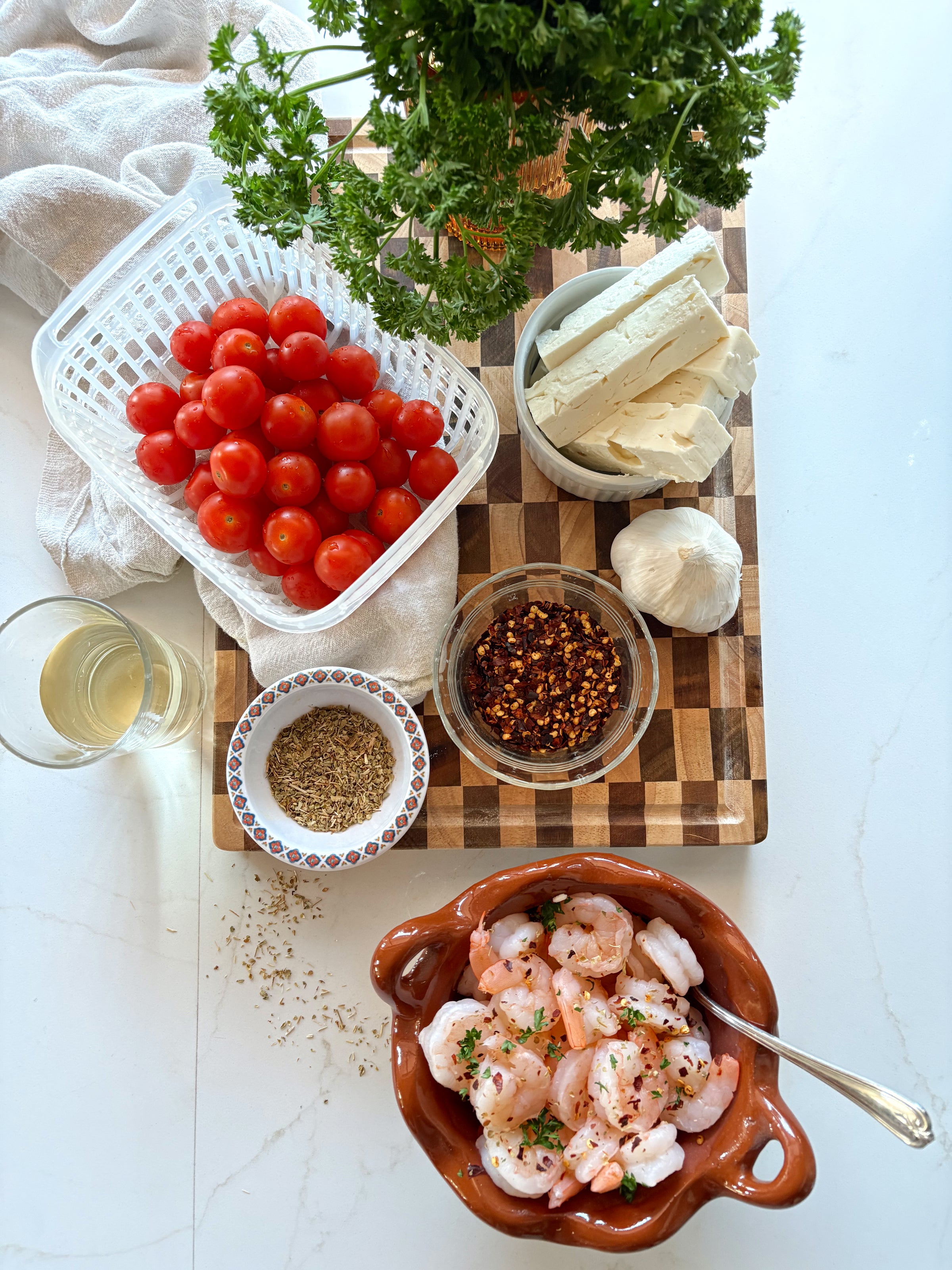A wooden board displays cherry tomatoes, feta cheese, fresh parsley, garlic, dried herbs, crushed red pepper, and a glass of white wine. In front, a dish holds cooked shrimp garnished with herbs.